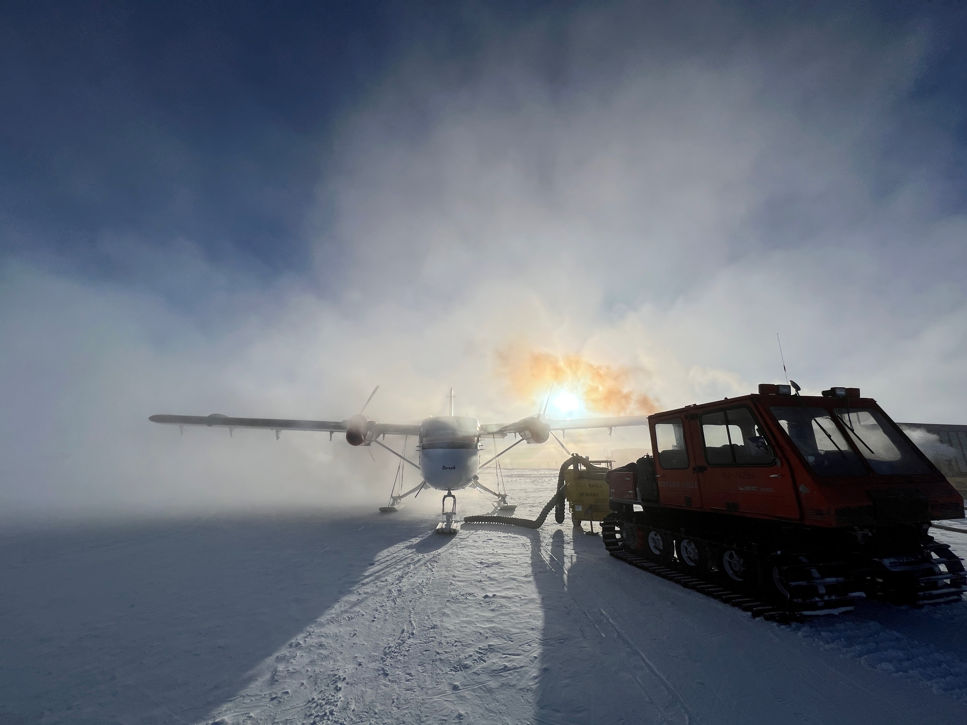 Mist shrouds small prop plane is refueled with snowcat vehicle in foreground