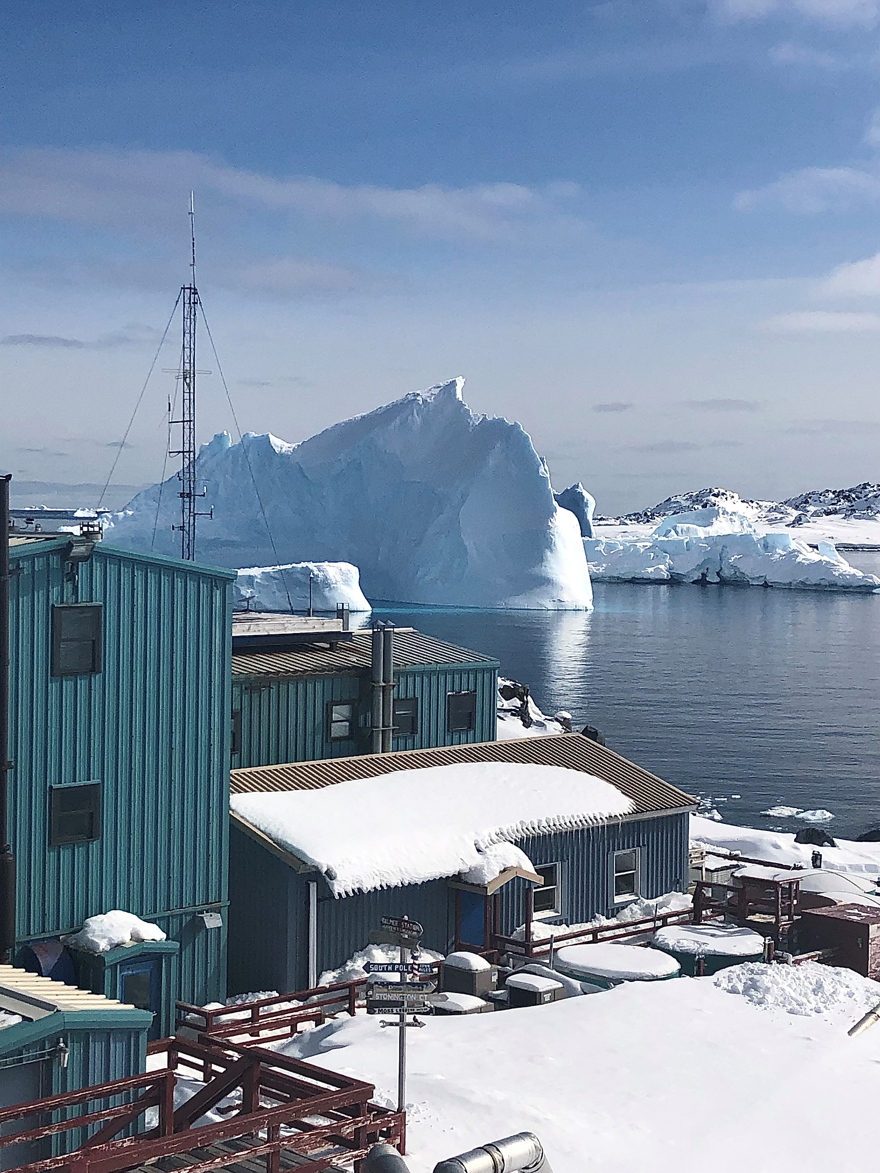 A green building with snow-covered roof with still water and iceberg in background