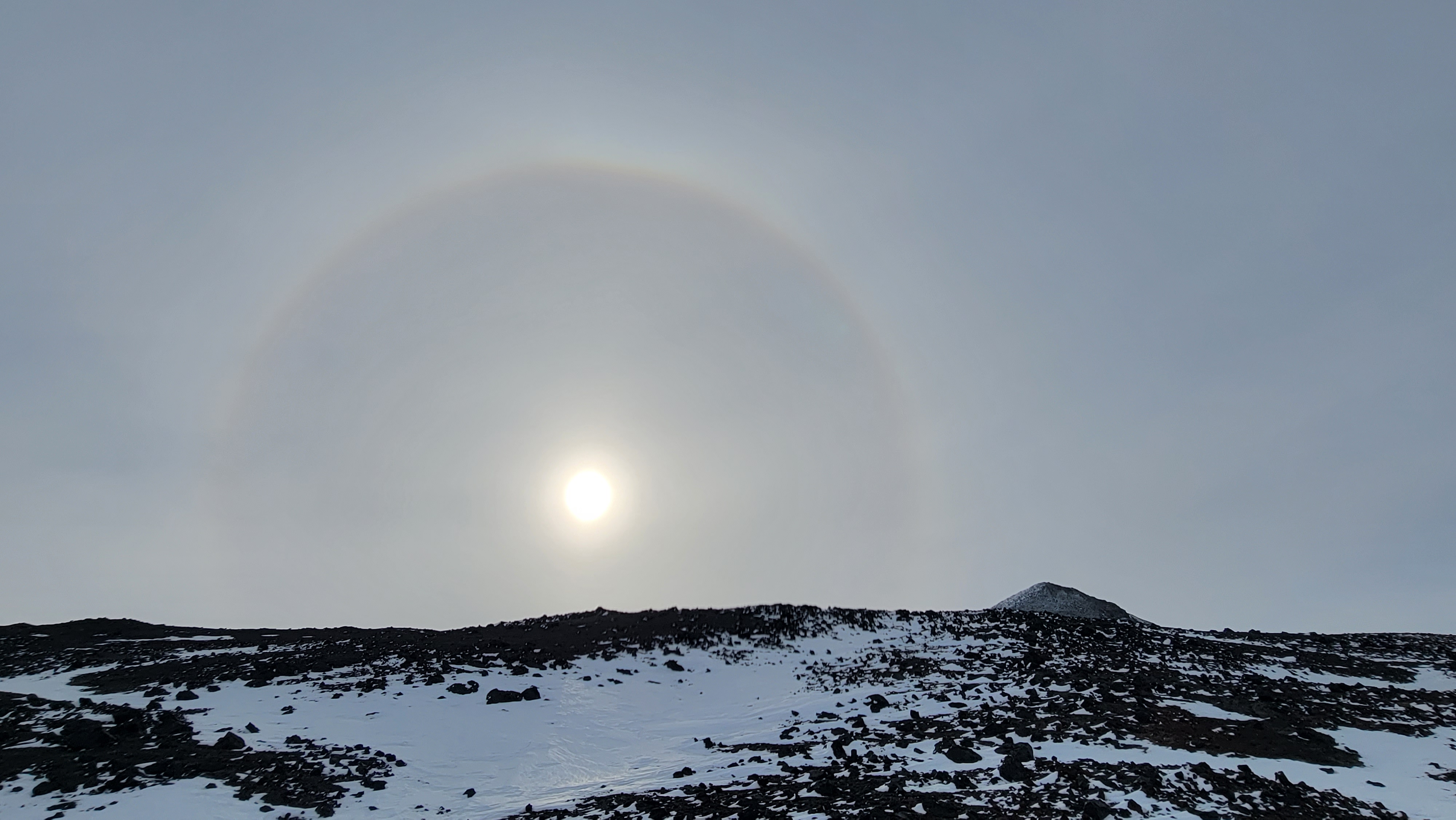 A halo-sun rises above a rocky and snowy hilltop