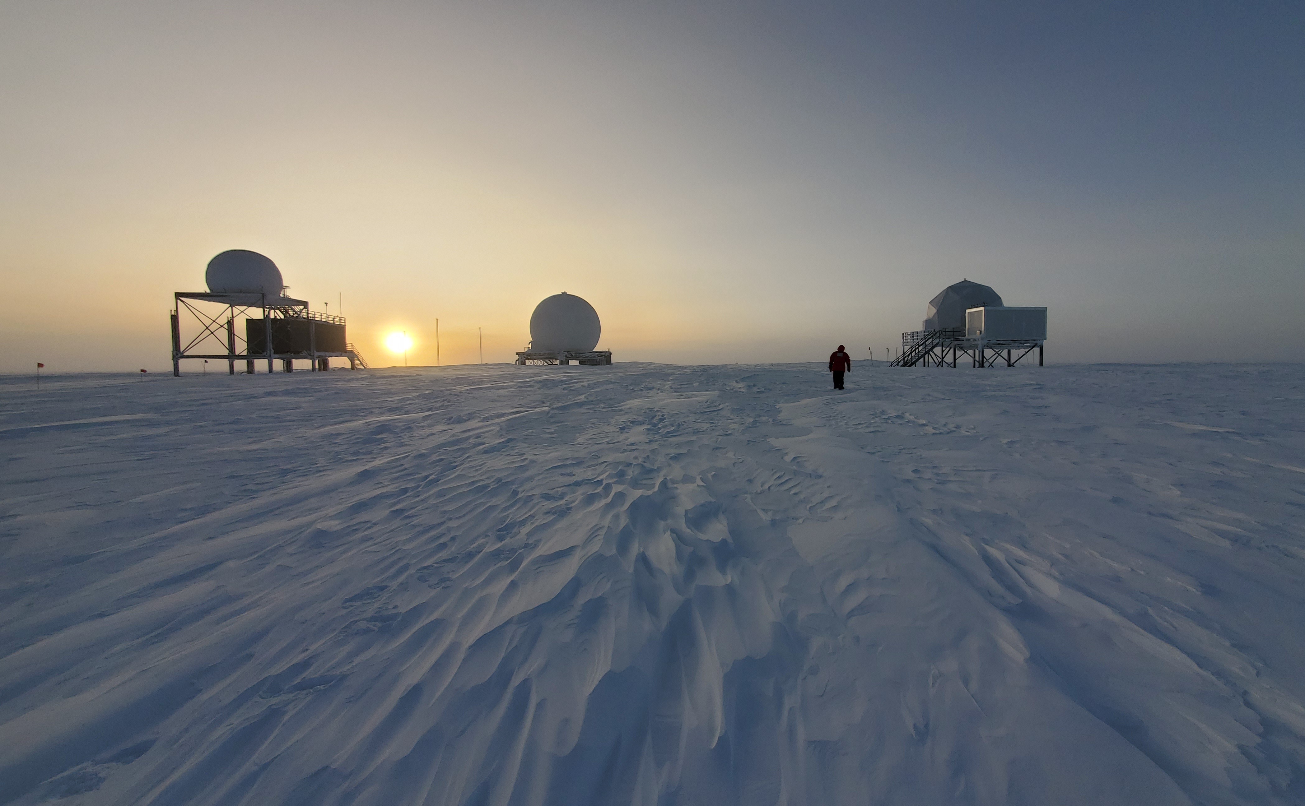 A snowy landscape with buildings and structures in distance and sun rising on the horizon