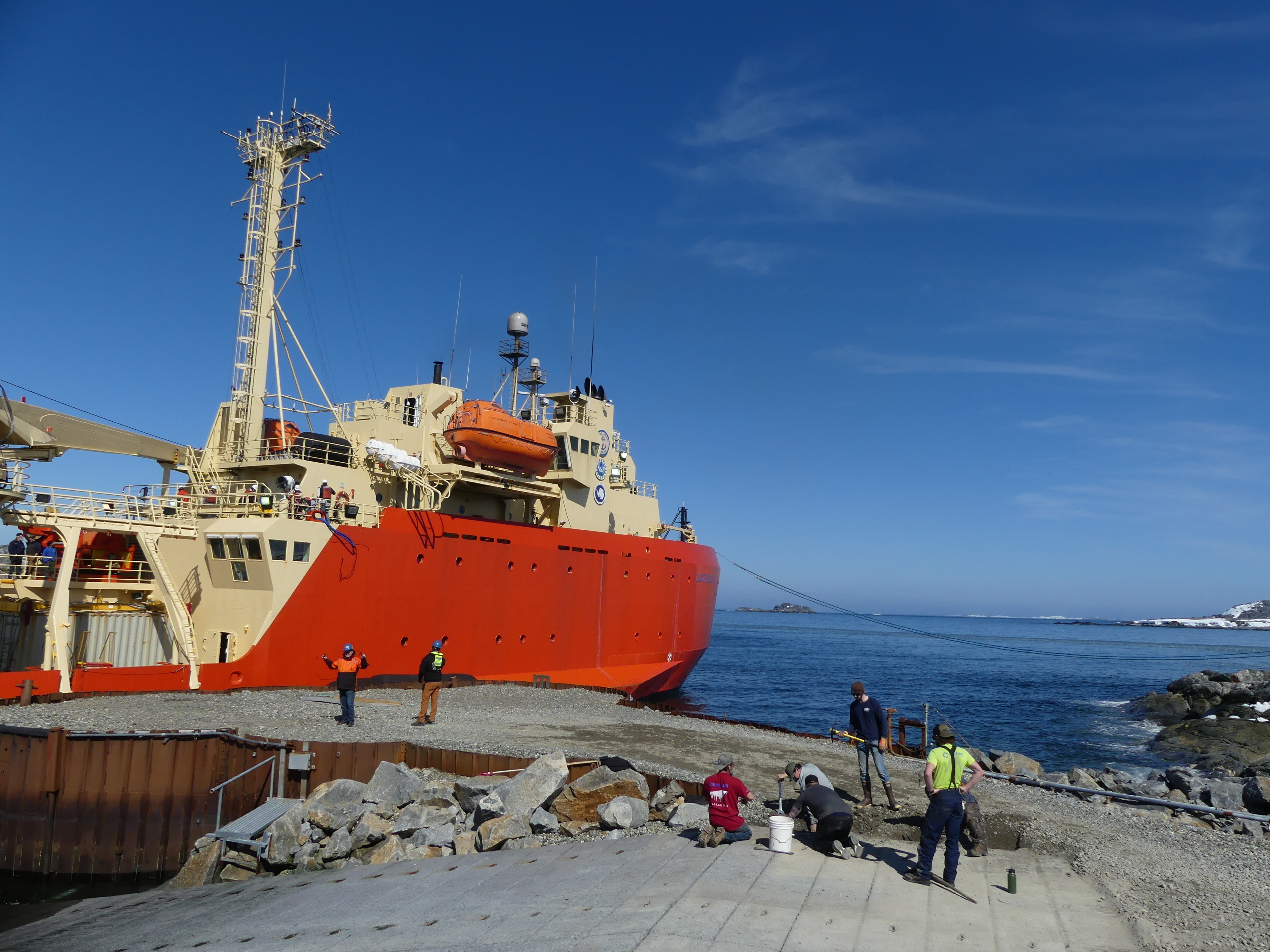 several people stand on a pier with a ship in the background.