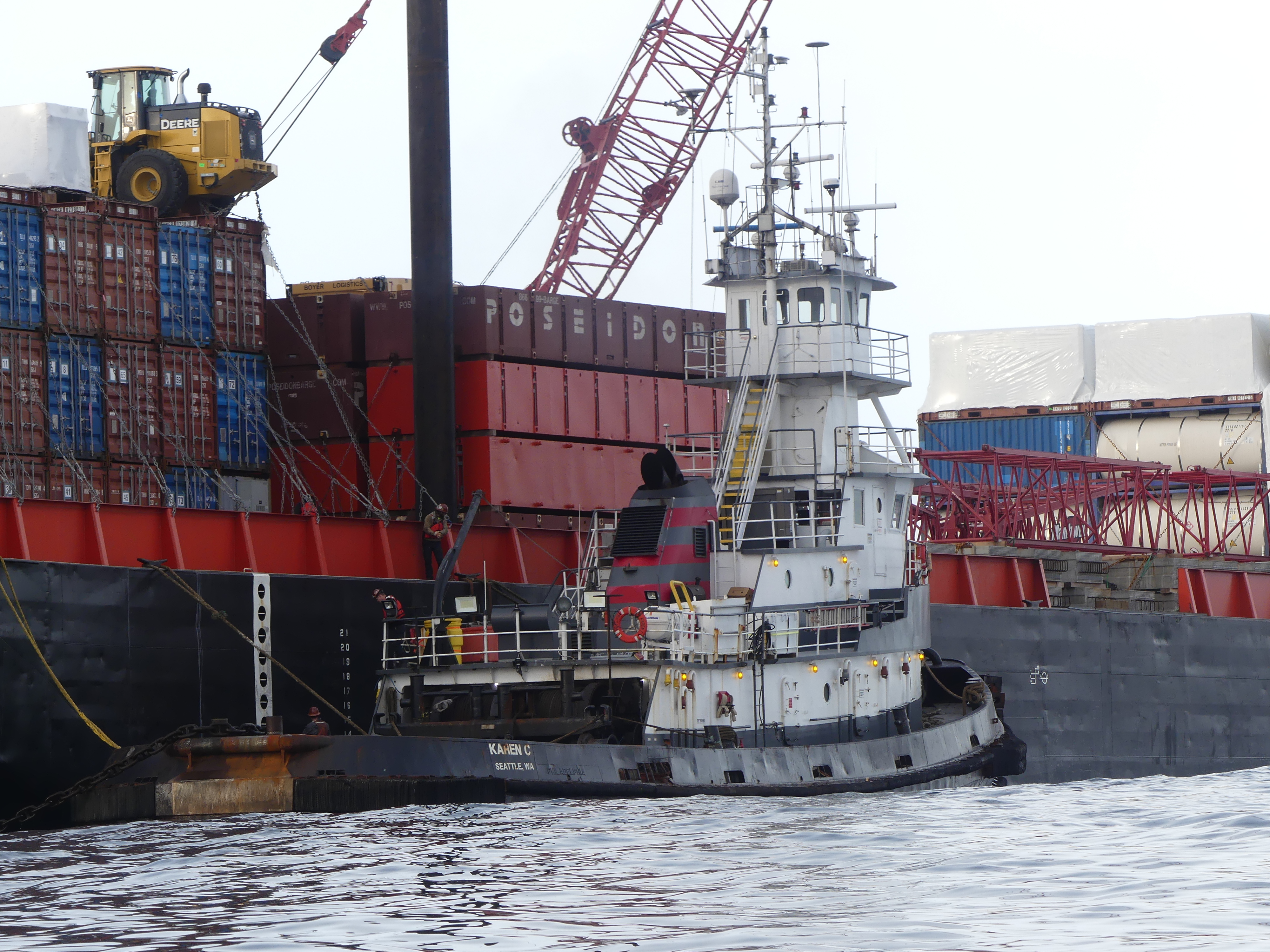 Close up view of a barge with lots of shipping containers and tugboat tied up next to it in the water. 