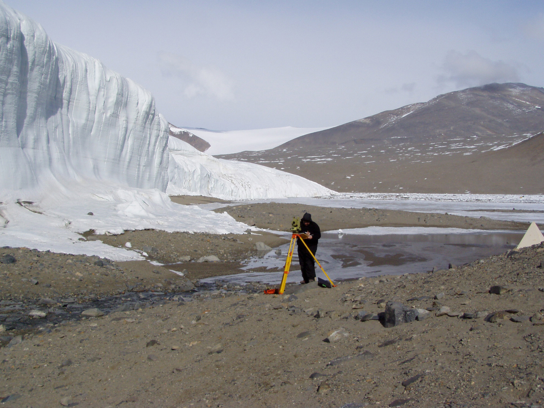 A person surveys an area with a transit, near a glacier and a stream.