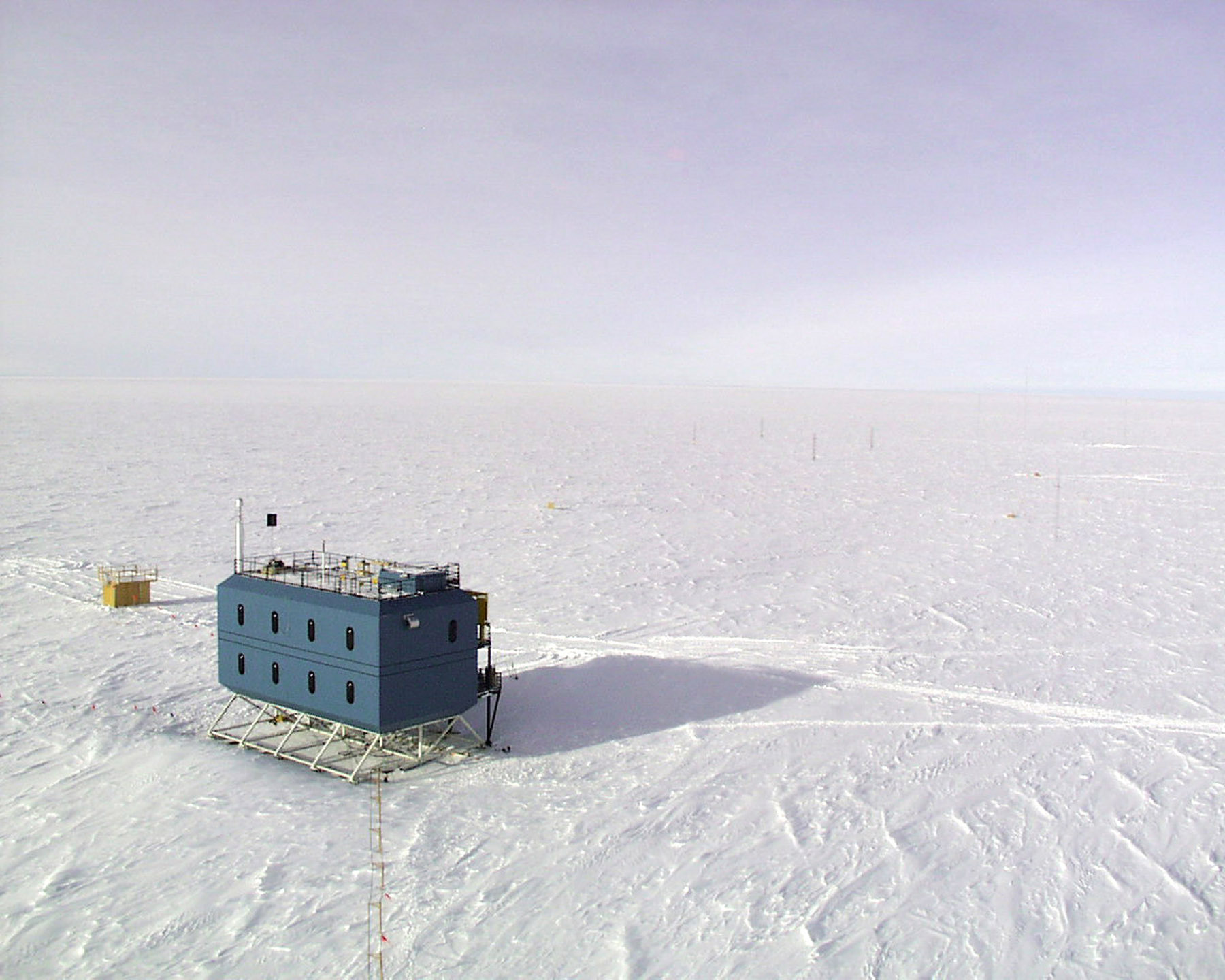 Aerial view of a blue building on stilts sitting on a vast expanse of white snow.