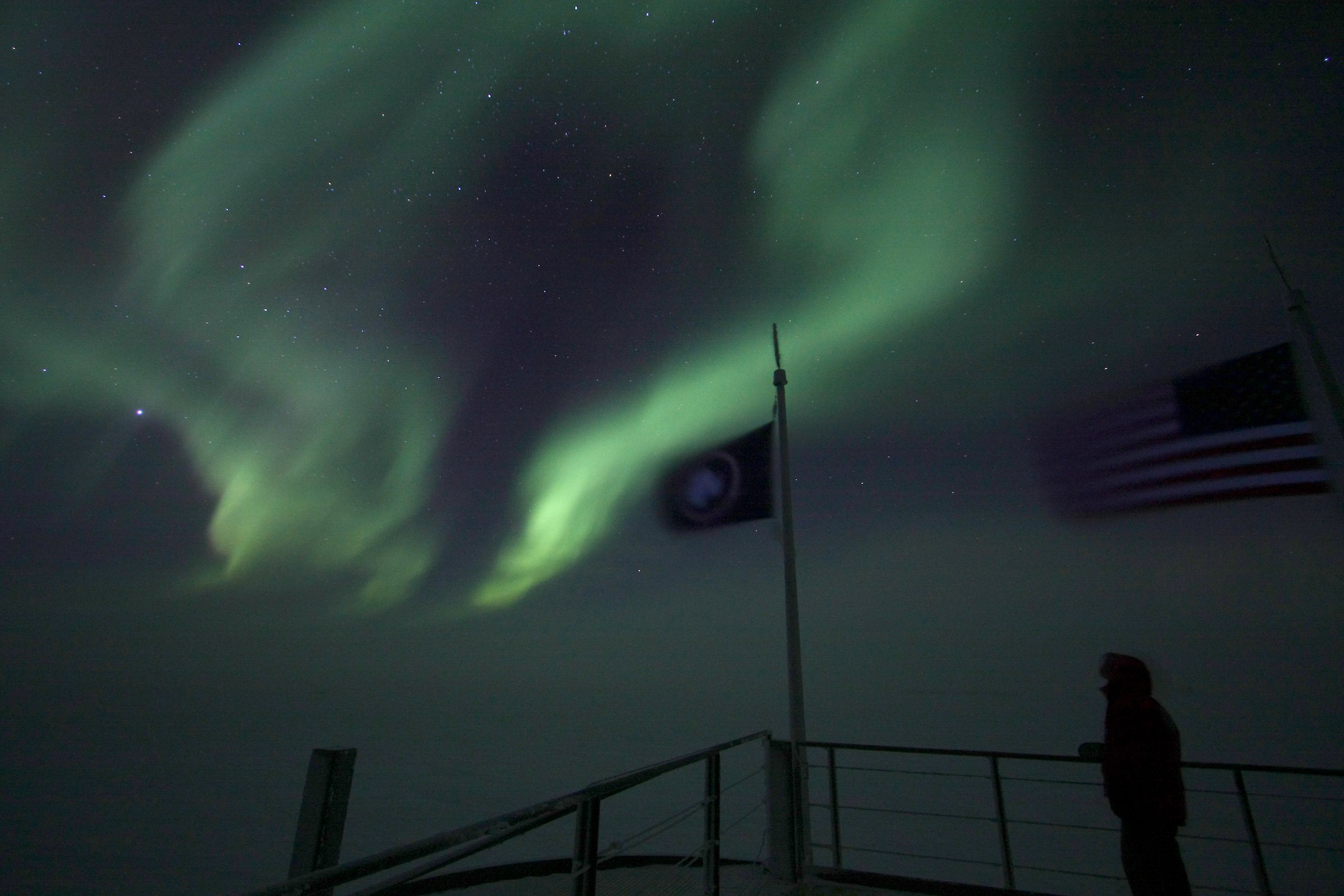 Auroras shimmer above a person and flags.