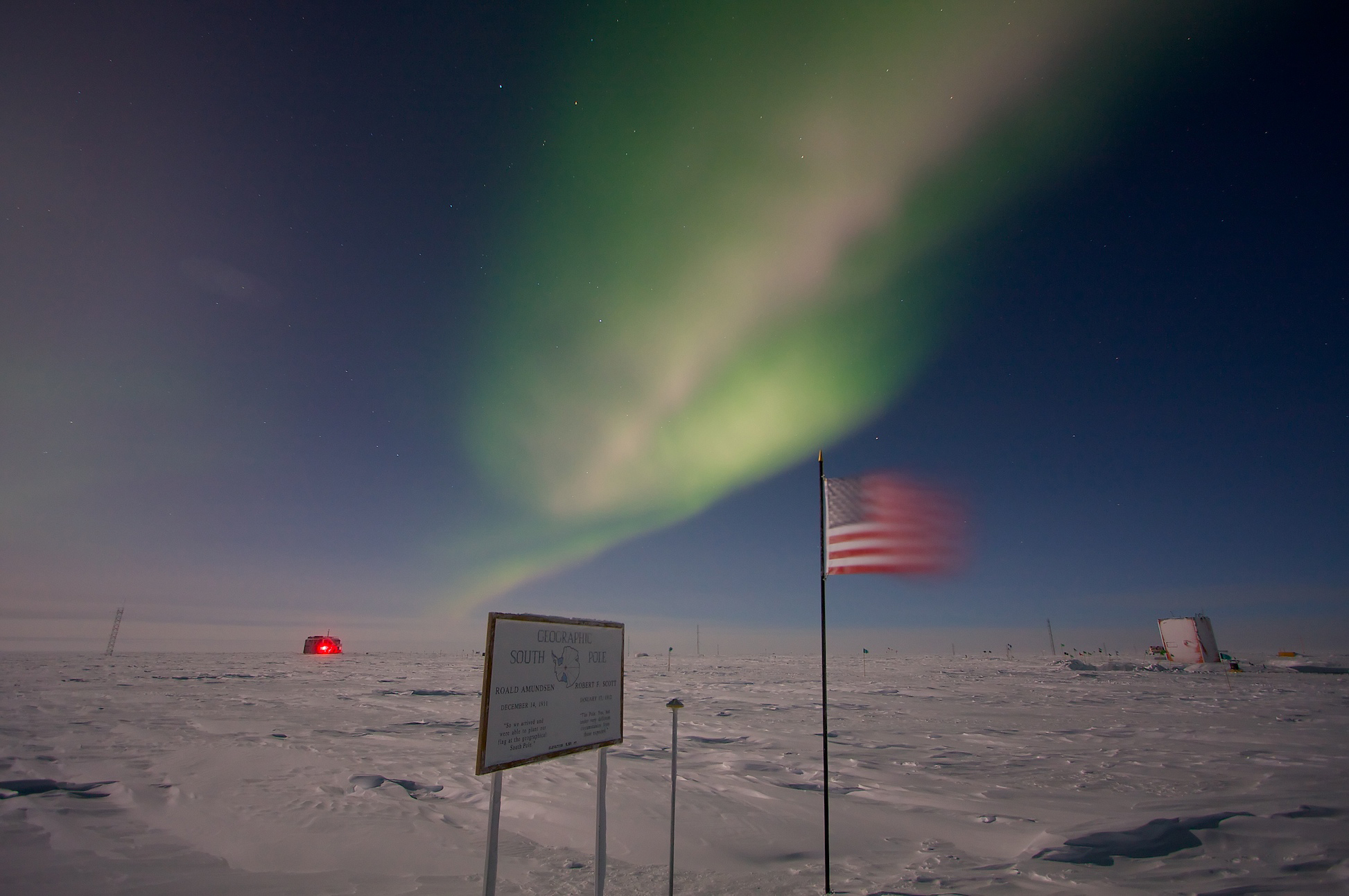 An aurora appears over a snowy landscape with a sign and flag in the foreground.