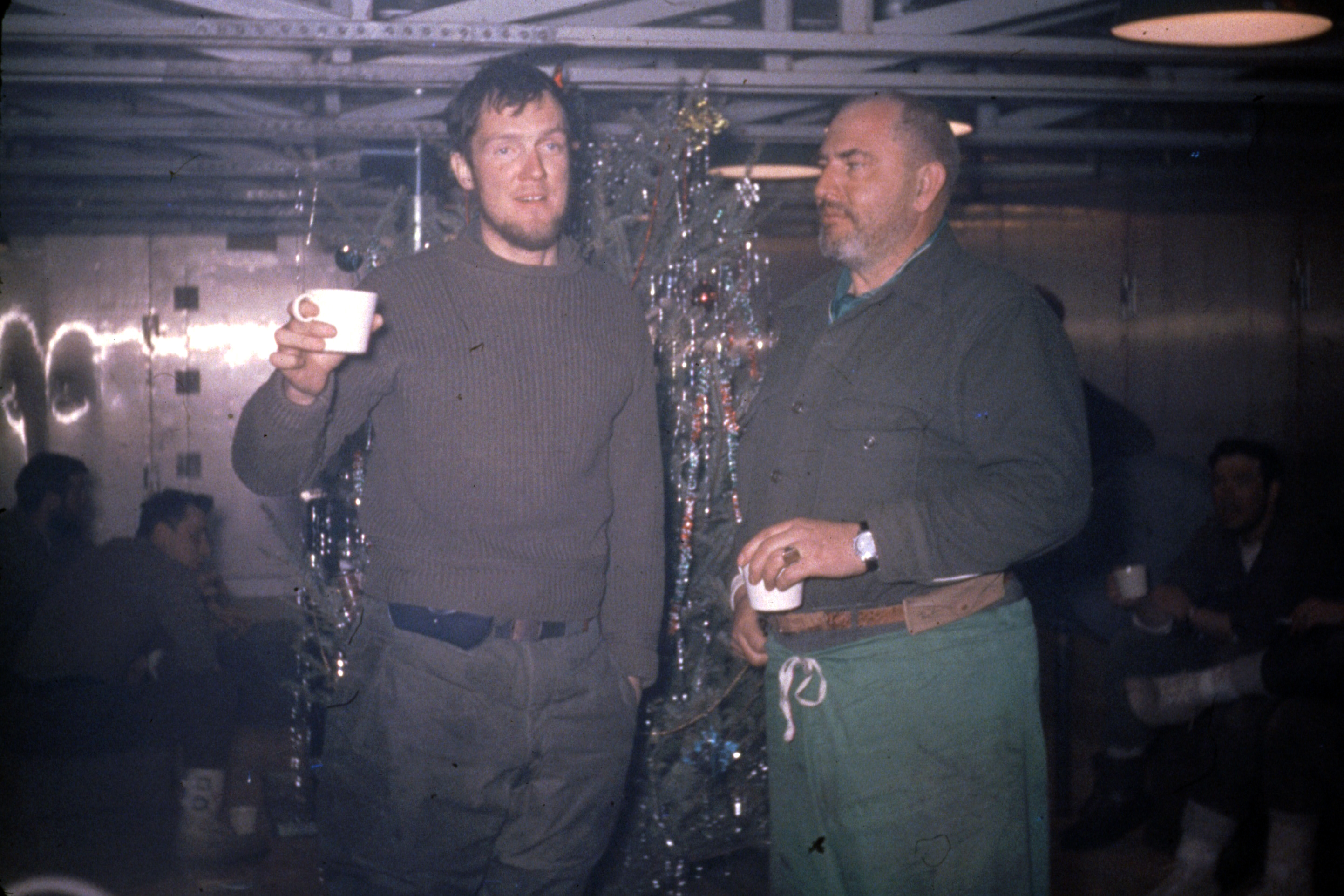 Two men share a drink in front of a Christmas tree.