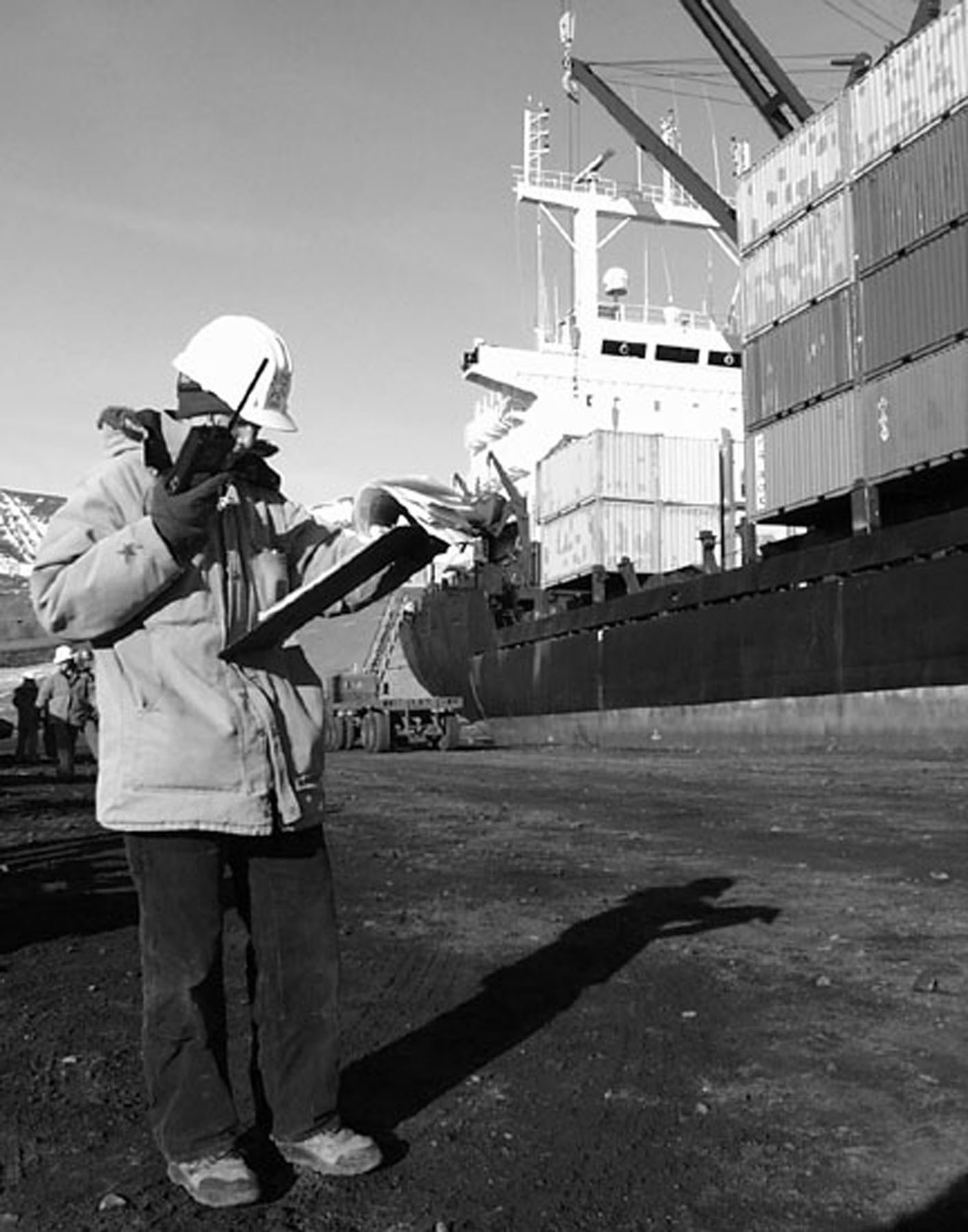 A person standing near a ship reads from a clipboard into a radio.