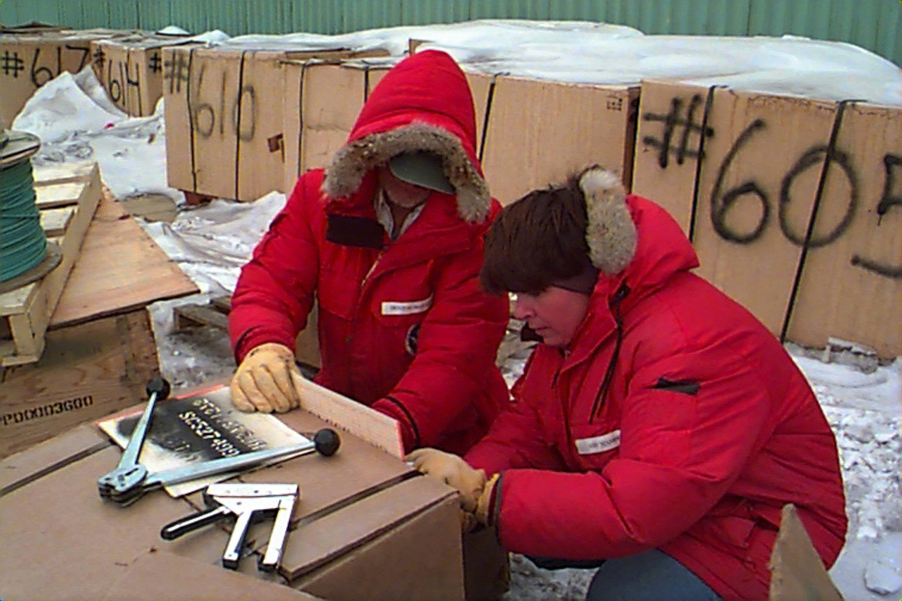 Two people in red parkas work with a large box.