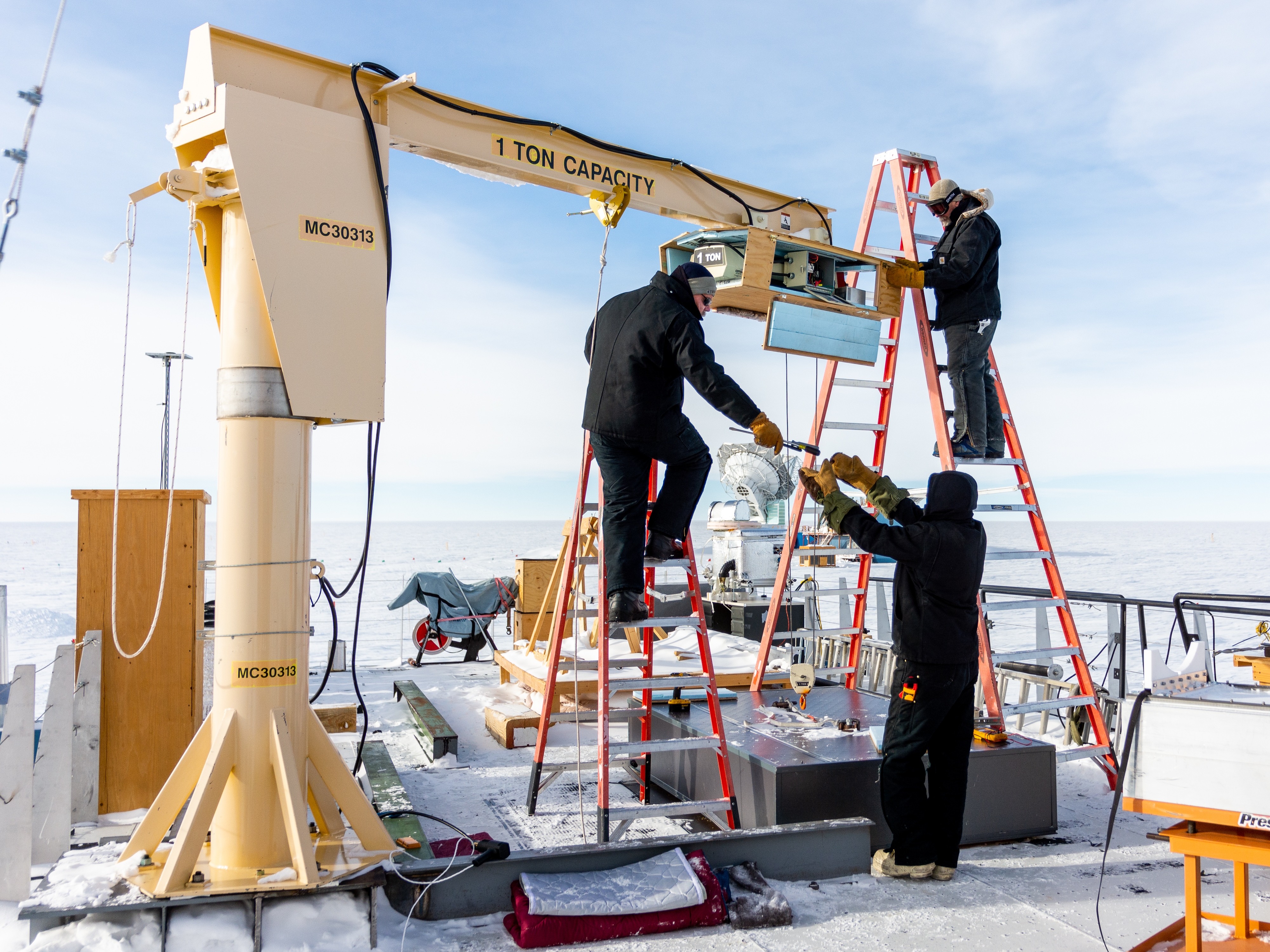 Workers work on a small crane with a snowy landscape in the background. 