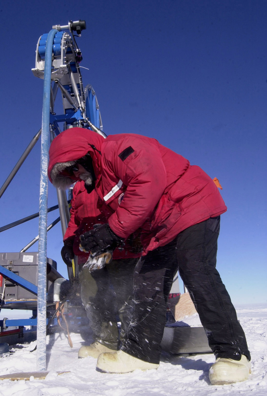 A man brushes snow off a drill that is coming up from below the ice. 