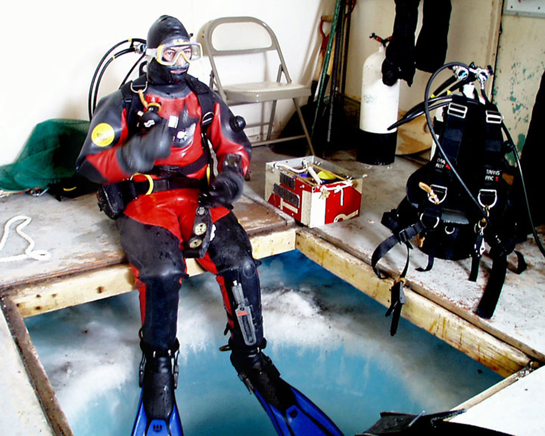 A SCUBA diver sits next to a hole in the ice.