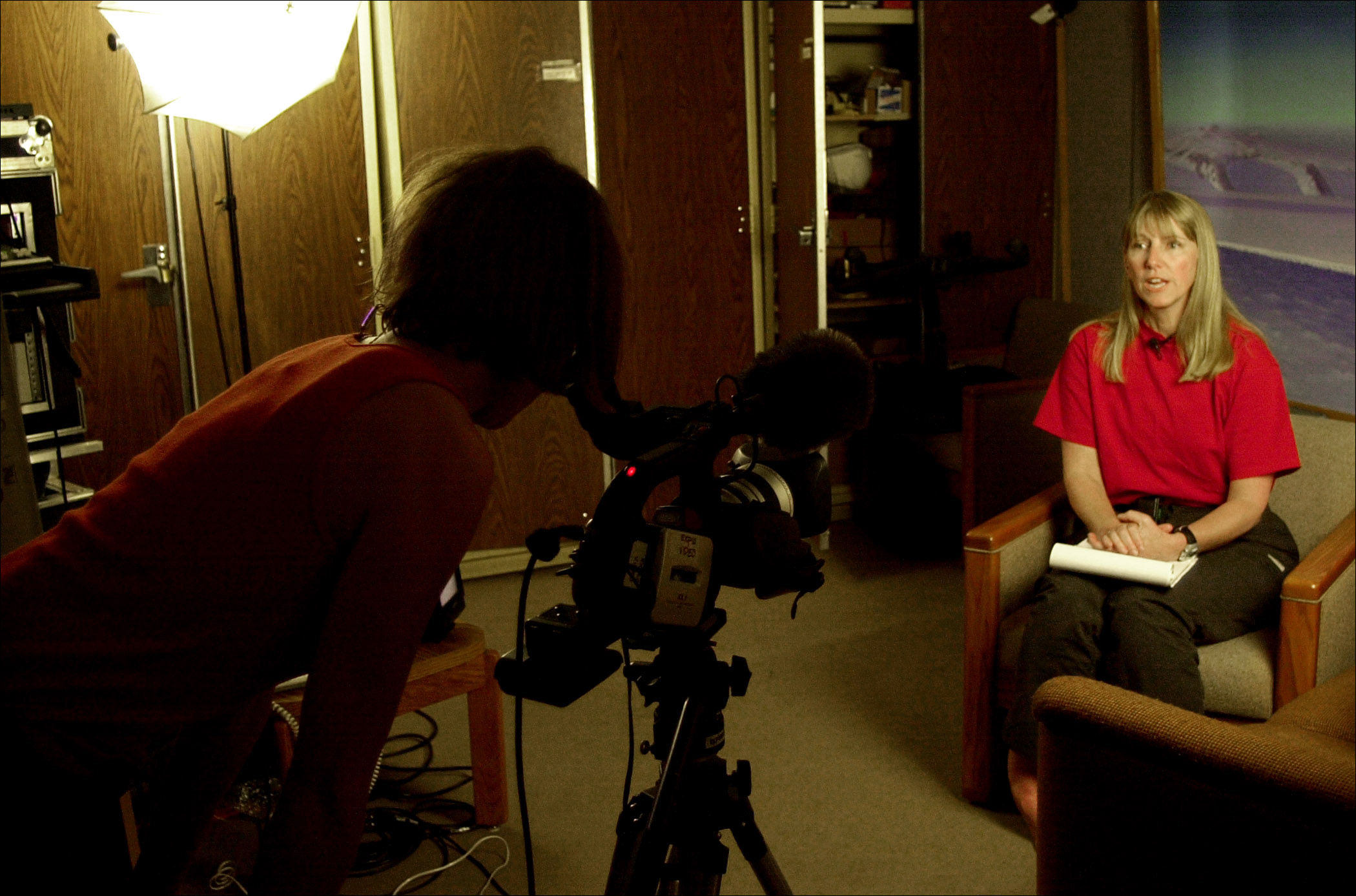 A blond-haired woman talks to a camera while another woman peers into the camera.