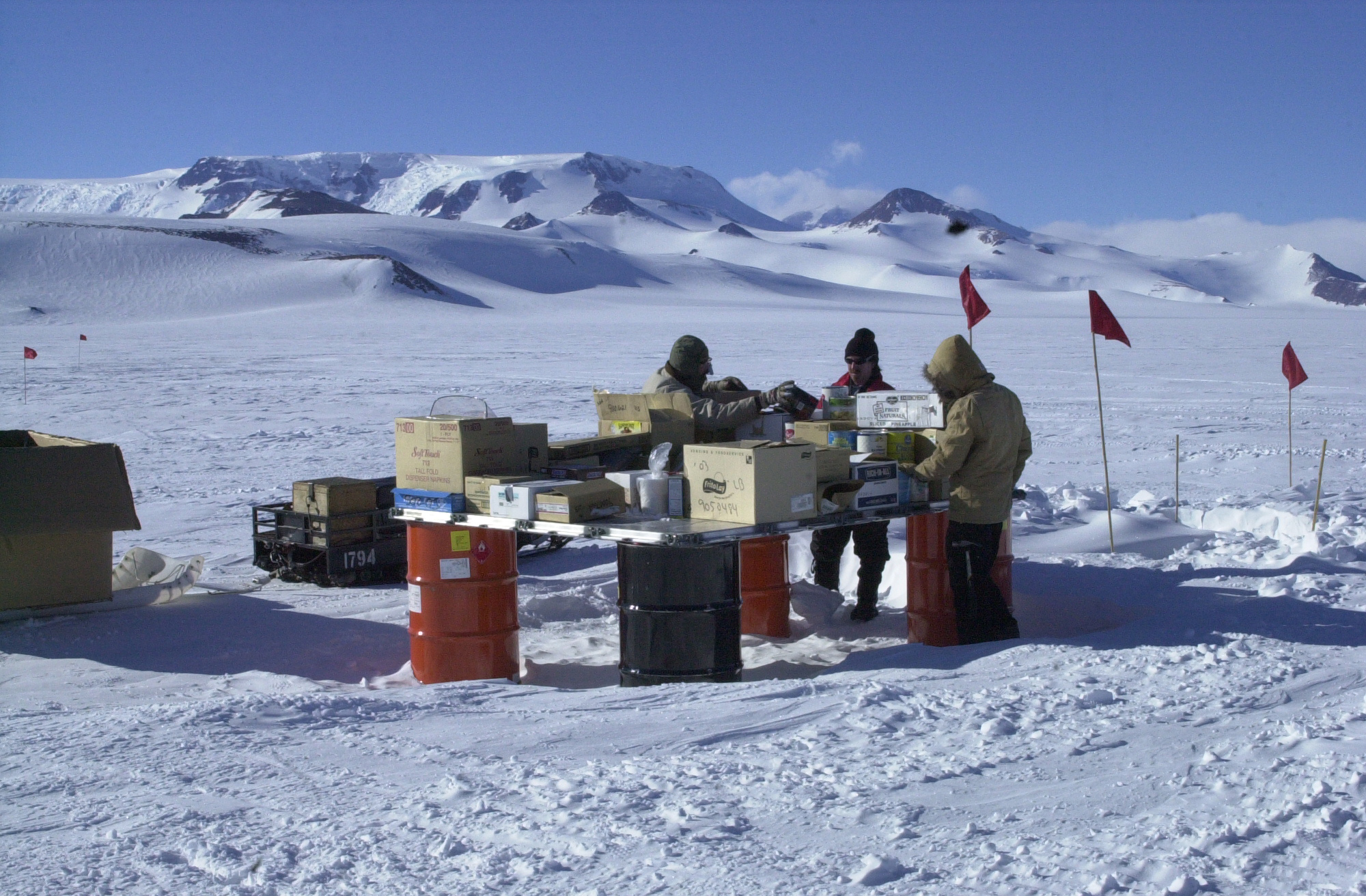 Three people stand around a table of food boxes outside in the snow.