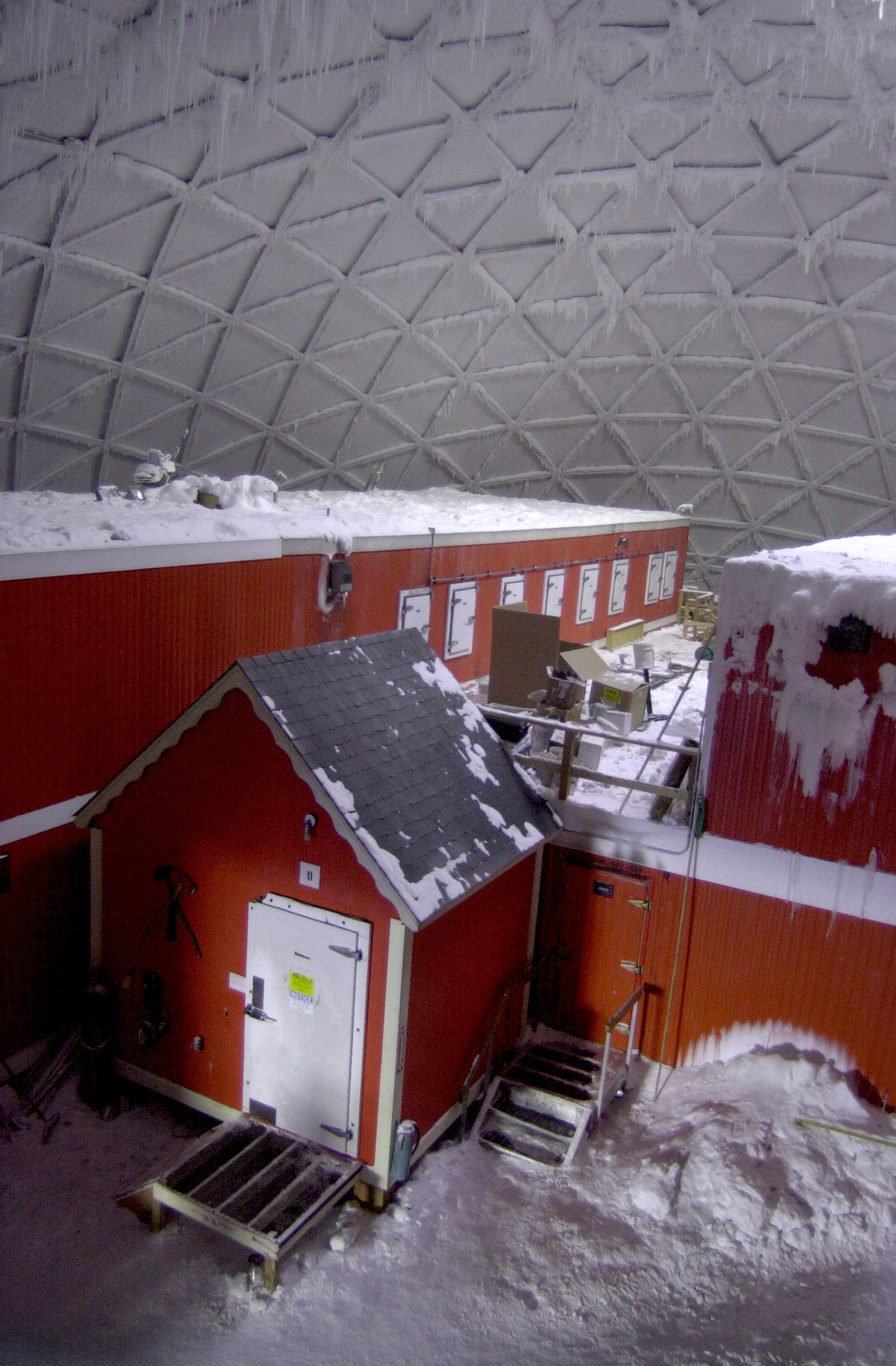 Elevated view inside geodesic dome looking down on three small red metal buildings covered with snow.