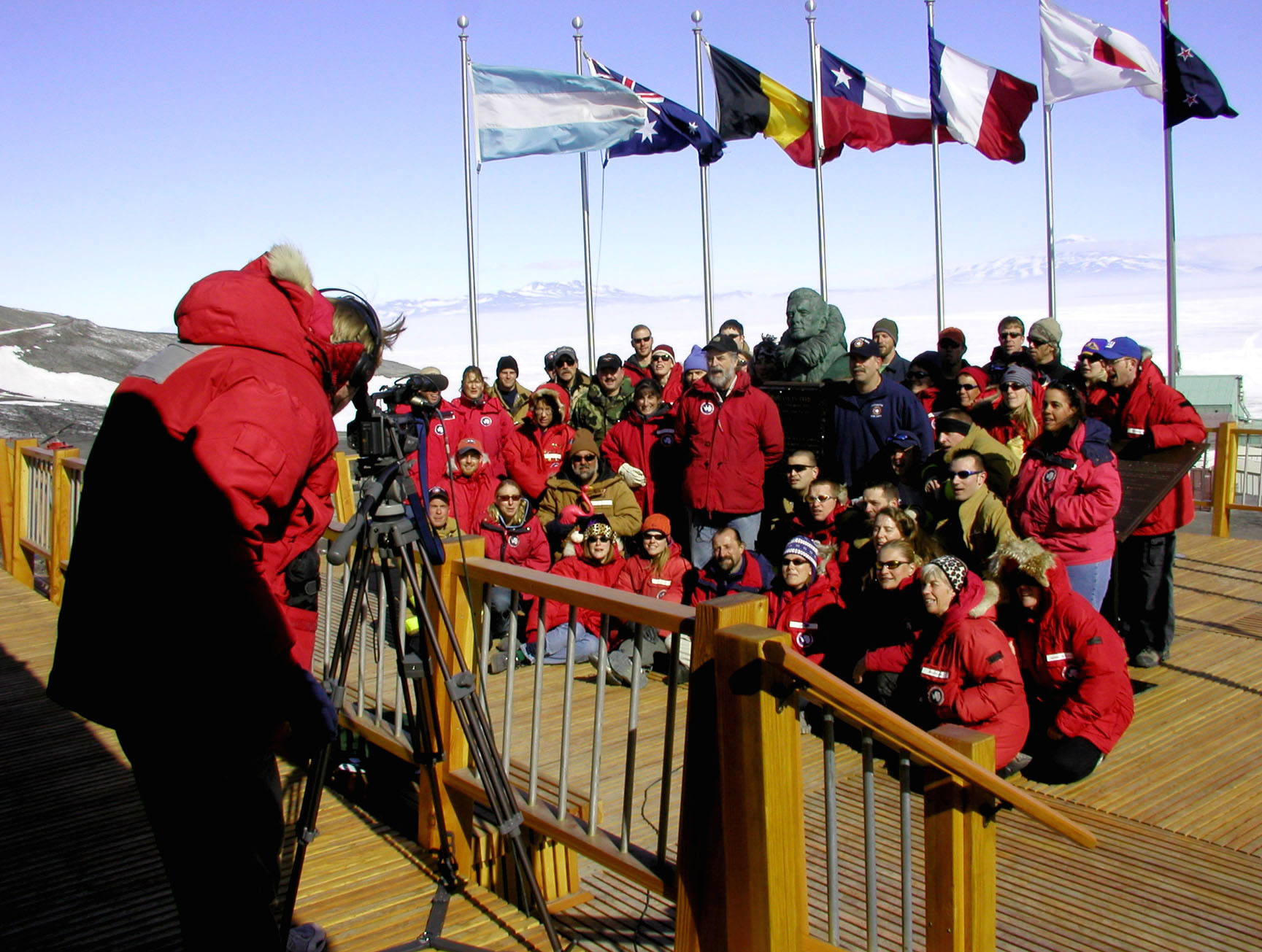 A man videotapes a group of about 40 people who are sitting outside in front of some flags.