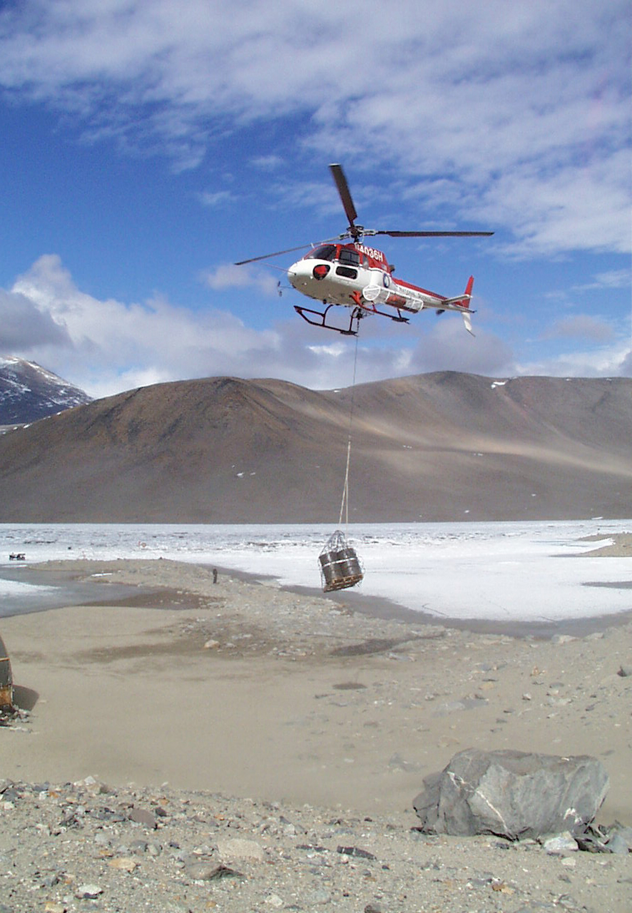 A helicopter carries a sling with cargo over a frozen lake.