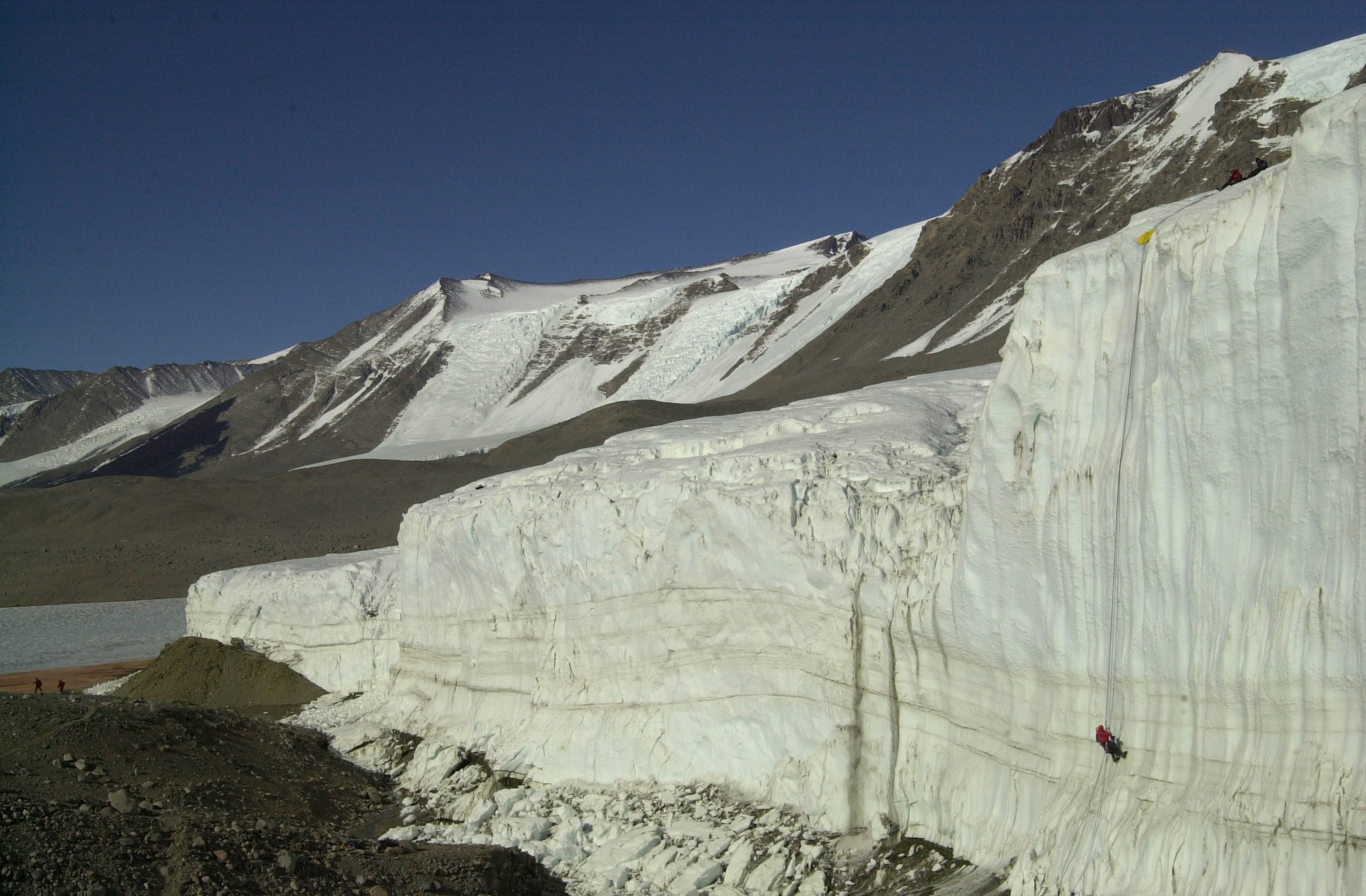 Two people climb up an enormous ice cliff with mountains in the background.