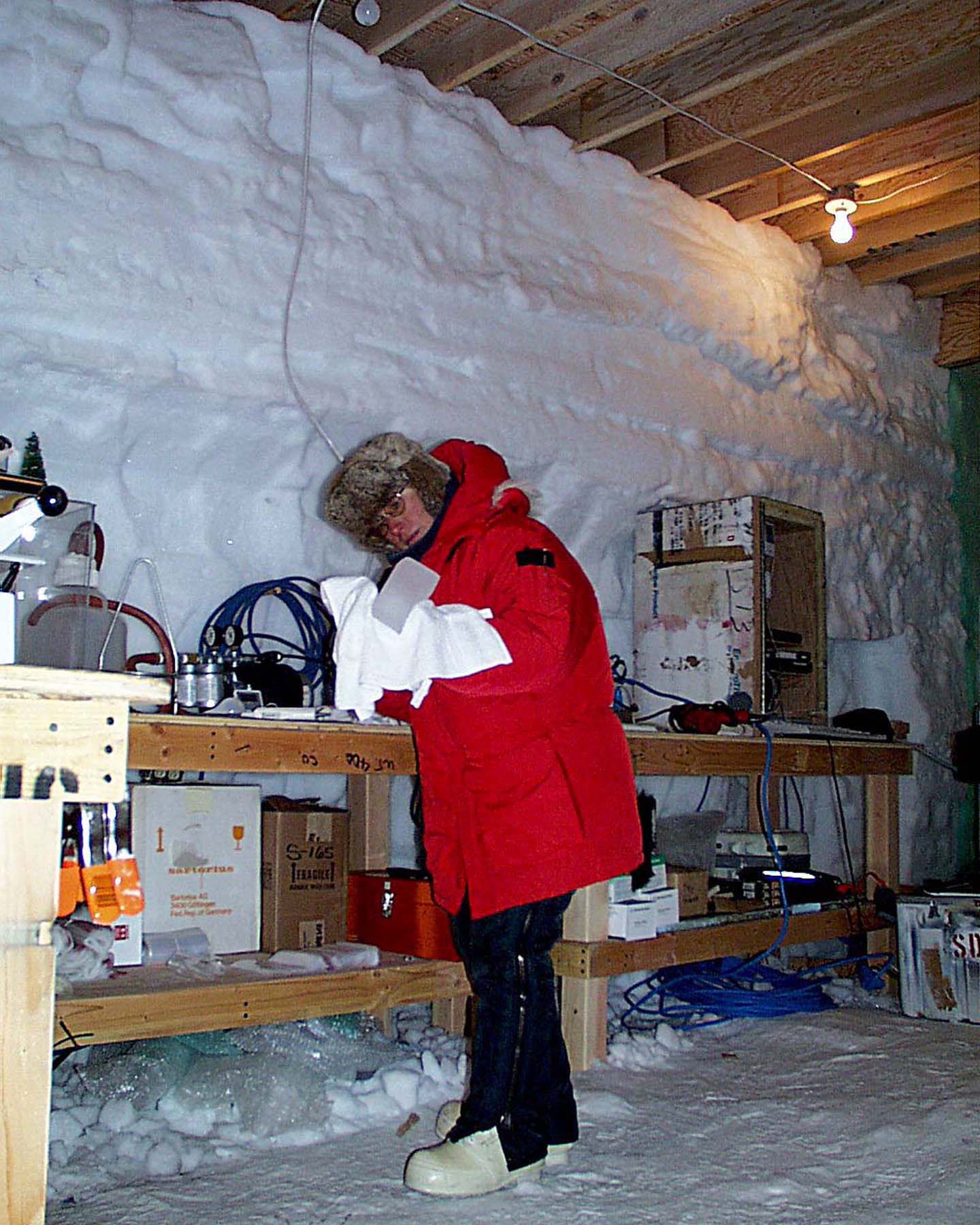 A man dressed in a red parka stands near a workbench.  