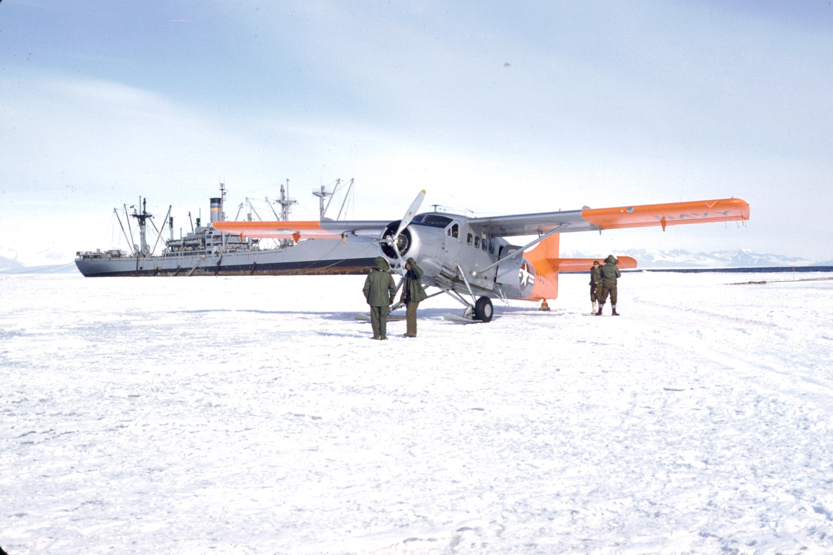 People stand around an airplane, with a ship in the distance.