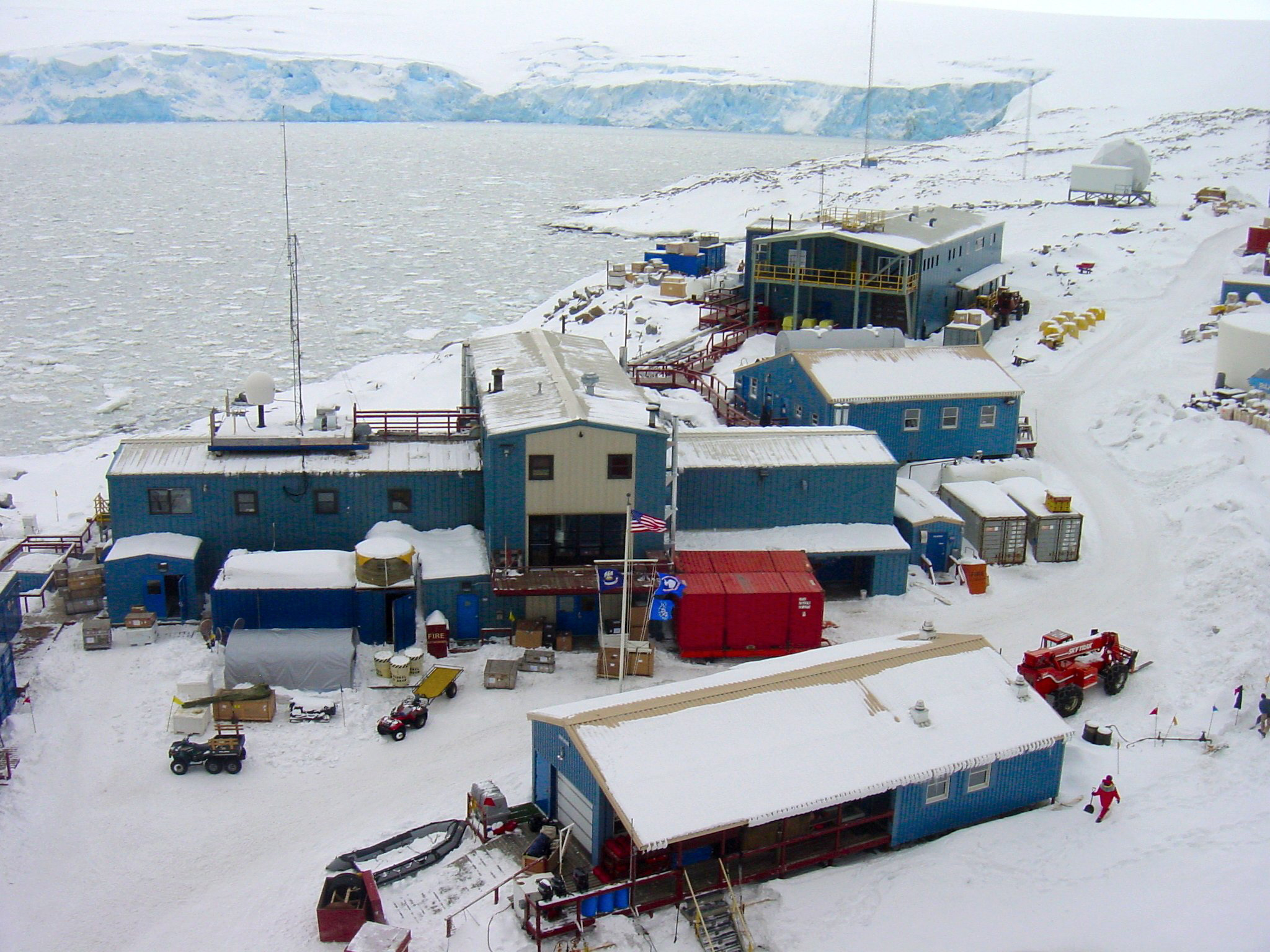 A cluster of blue buildings, in a snowy landscape, sit near open water.