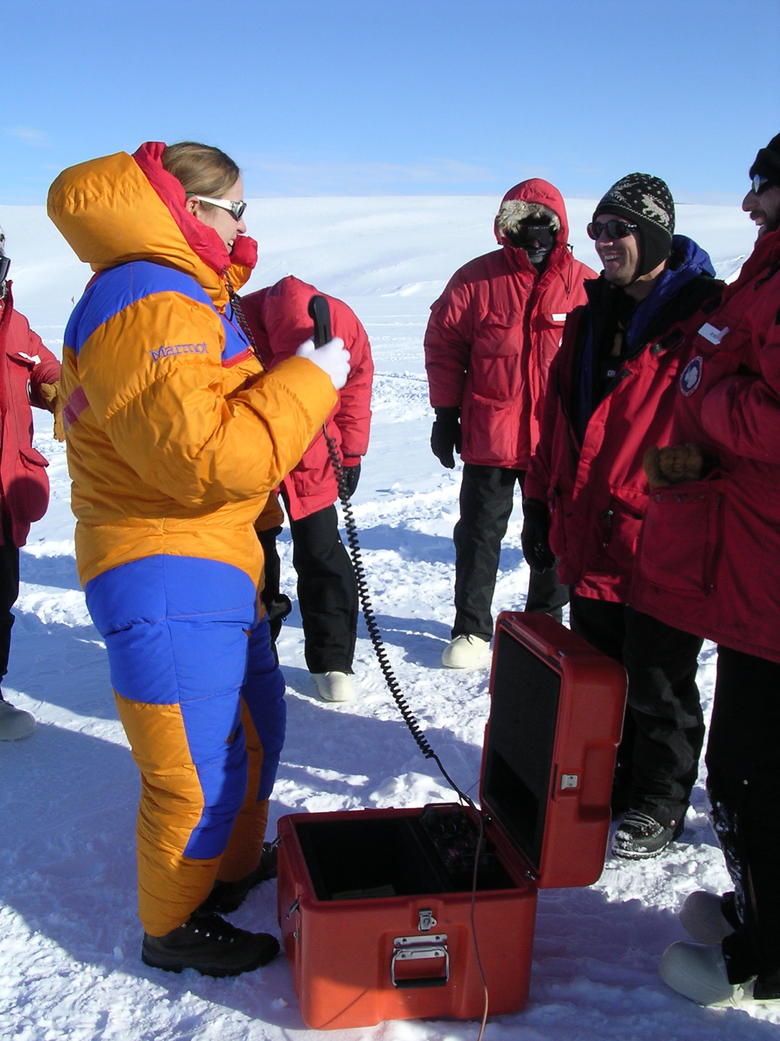 A woman holds a telephone-like object connected to a box on the ground. 