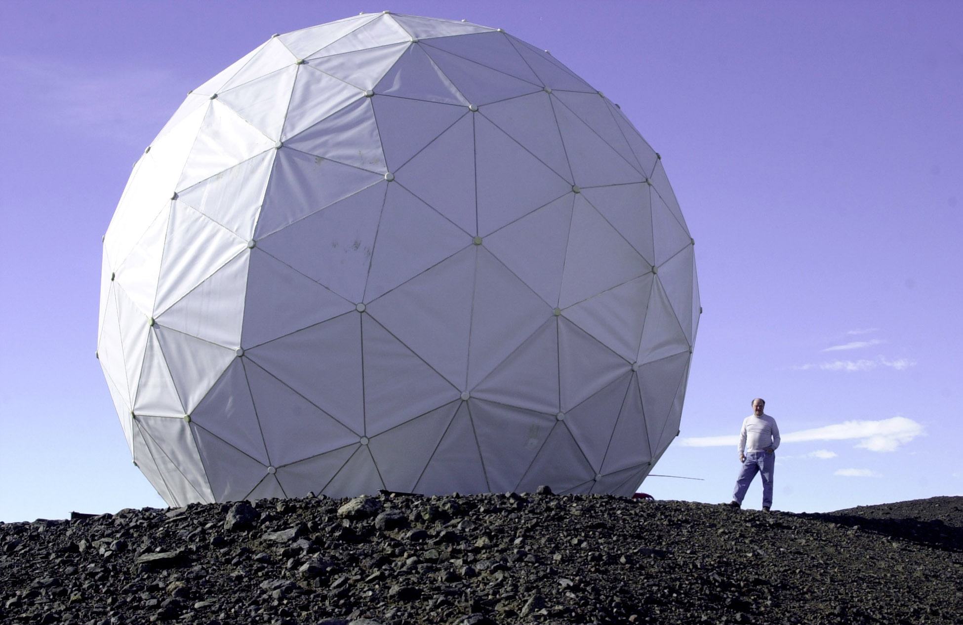 A man stands next to a large geodesic sphere on rocky ground.
