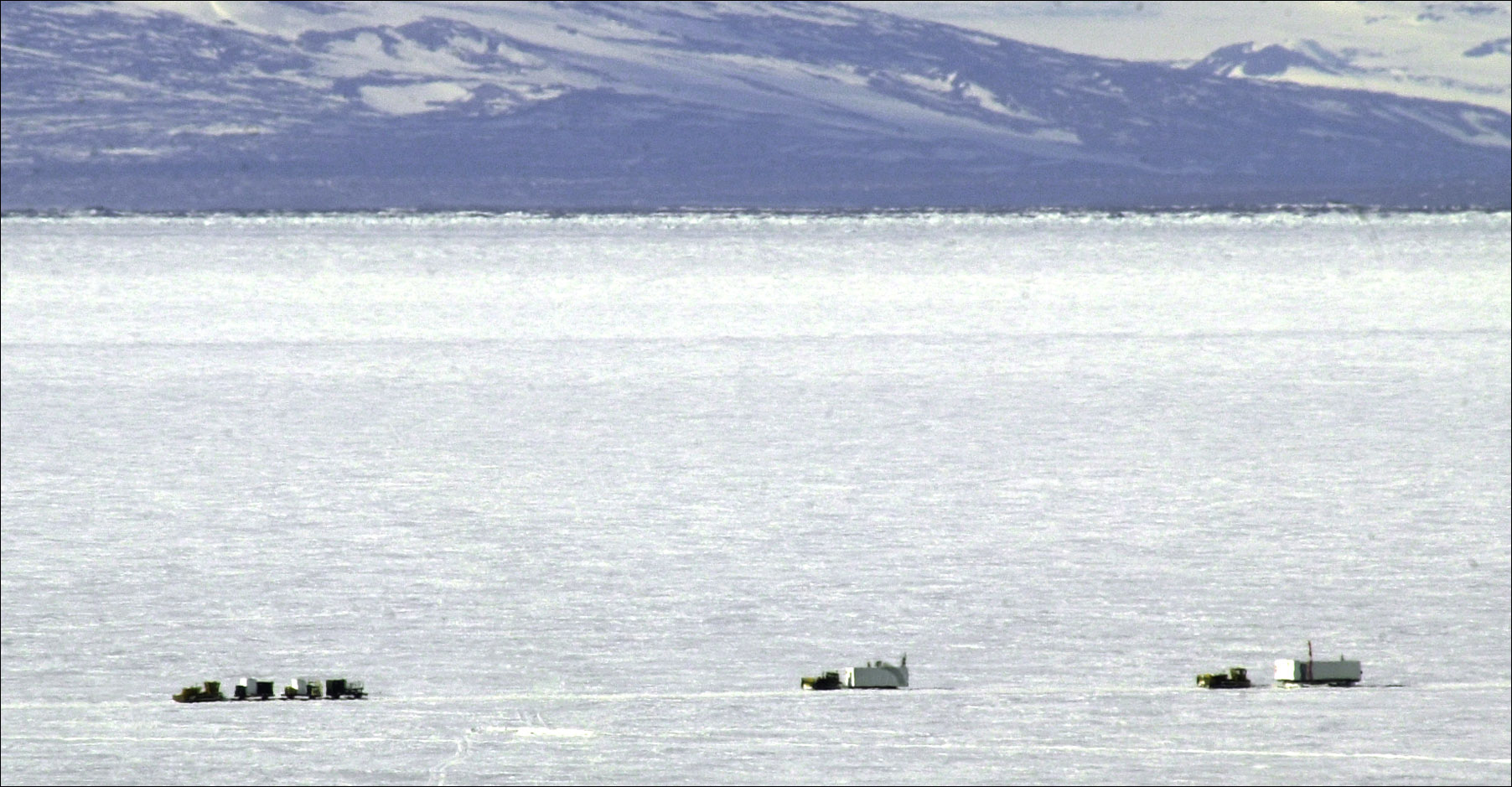 Distant shot of large tractors pulling buildings across white sea ice with mountains in the far distance.