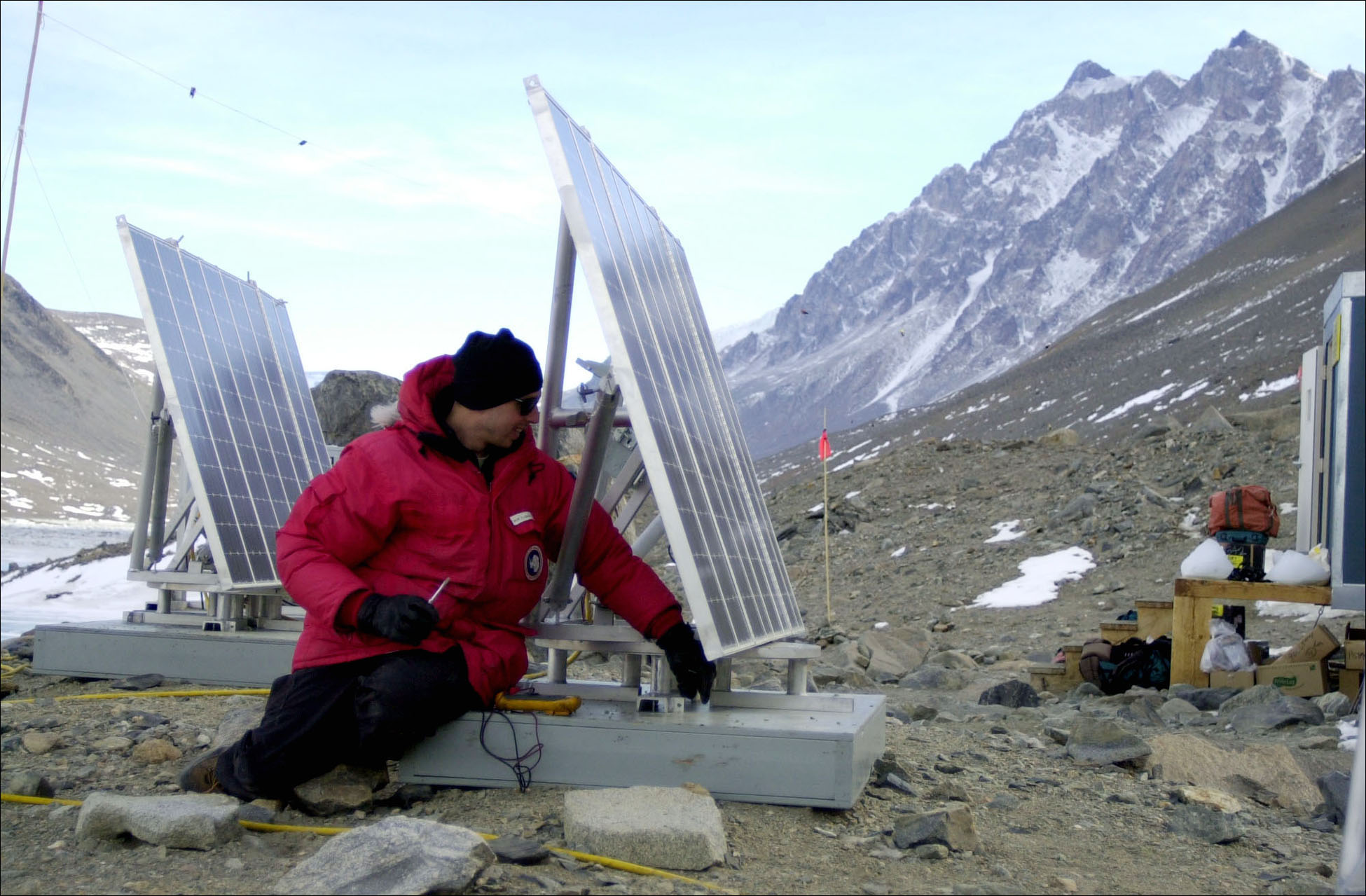 A man sits near two solar panels with mountains in the background. 