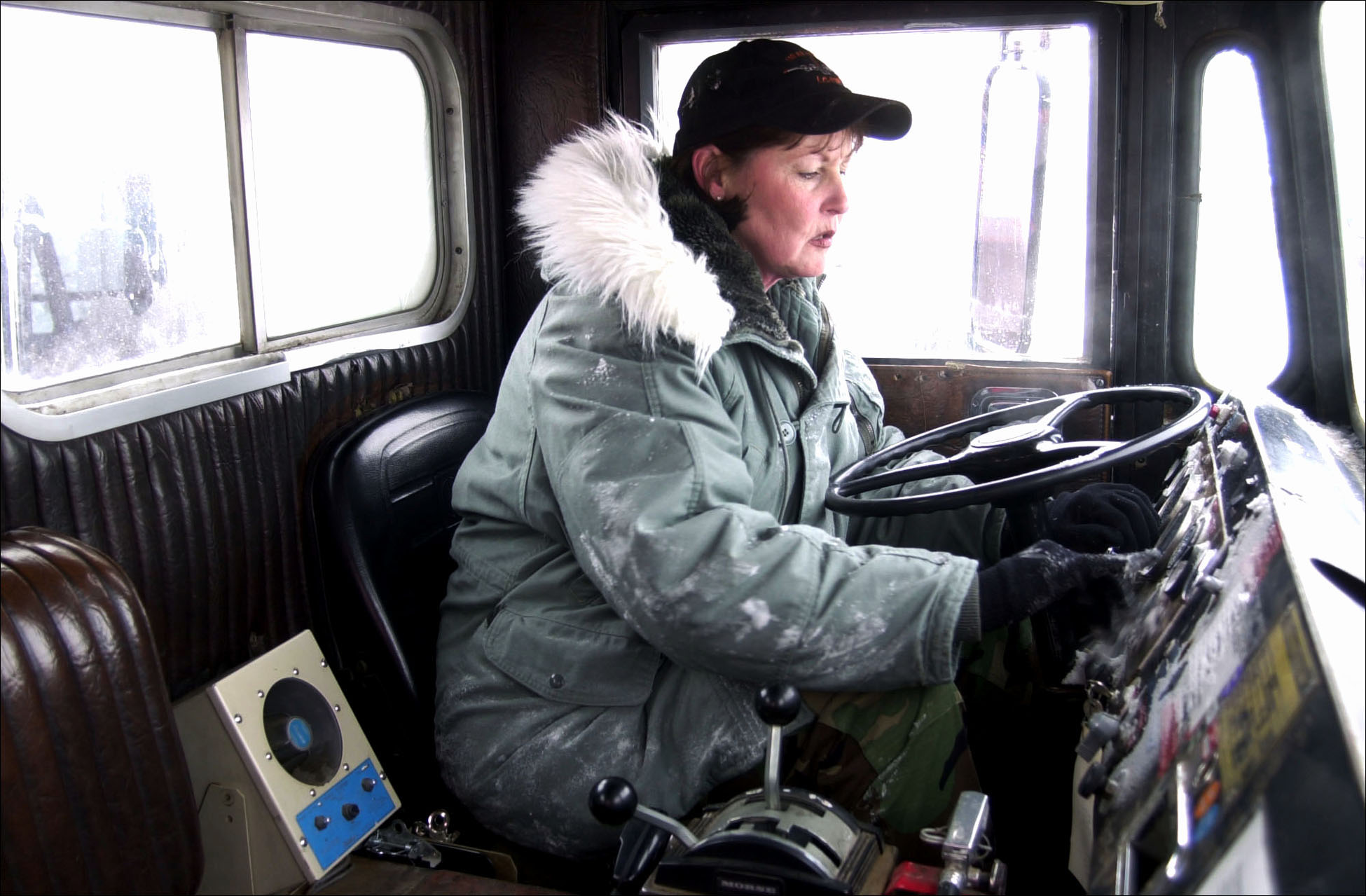 A woman wearing an army uniform and and black hat starts the engine of a large snow truck. 