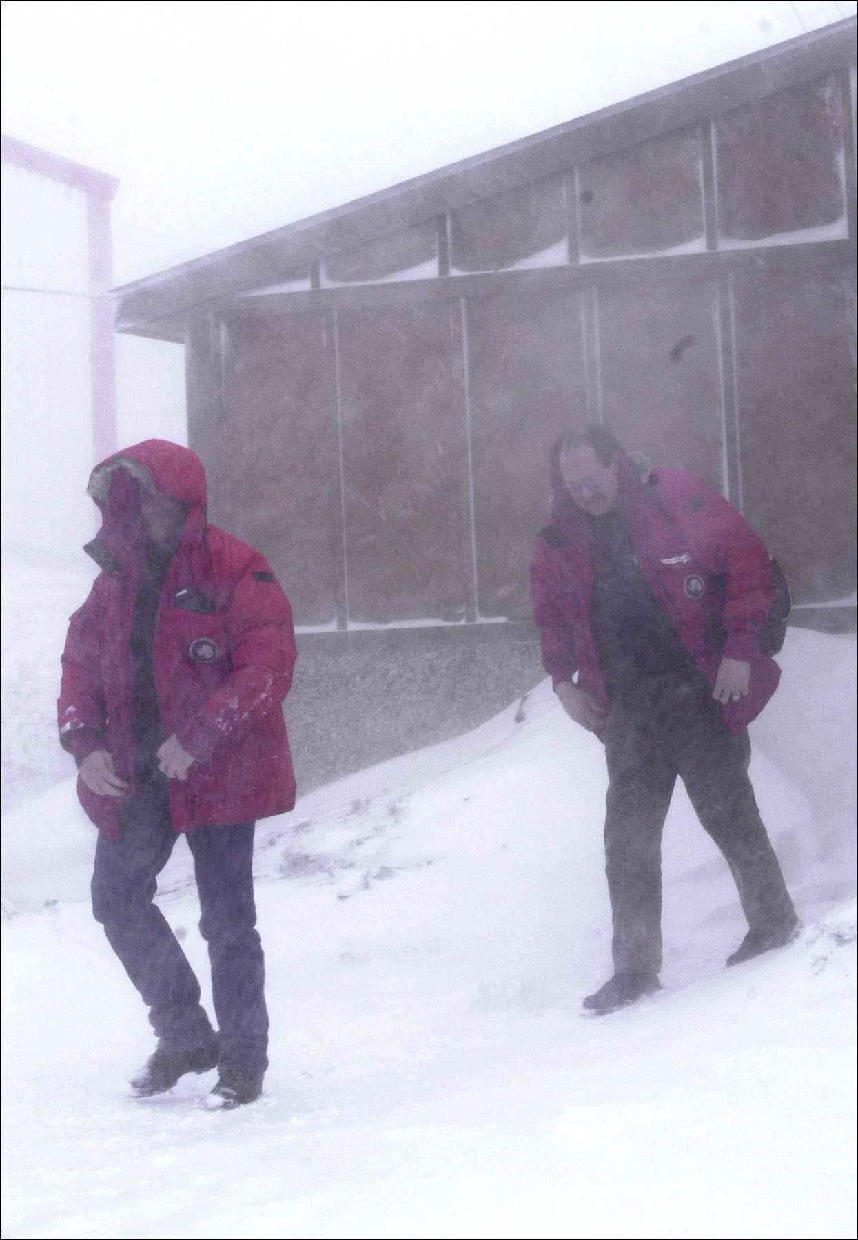 Two men in red parkas walk away from a building in a blizzard.