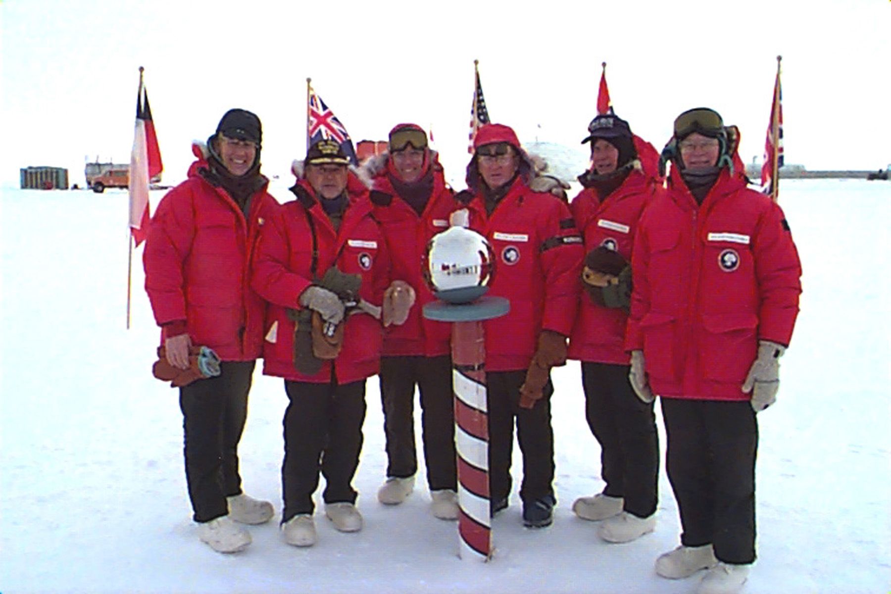 Six men in red parkas pose by a red and white striped pole.