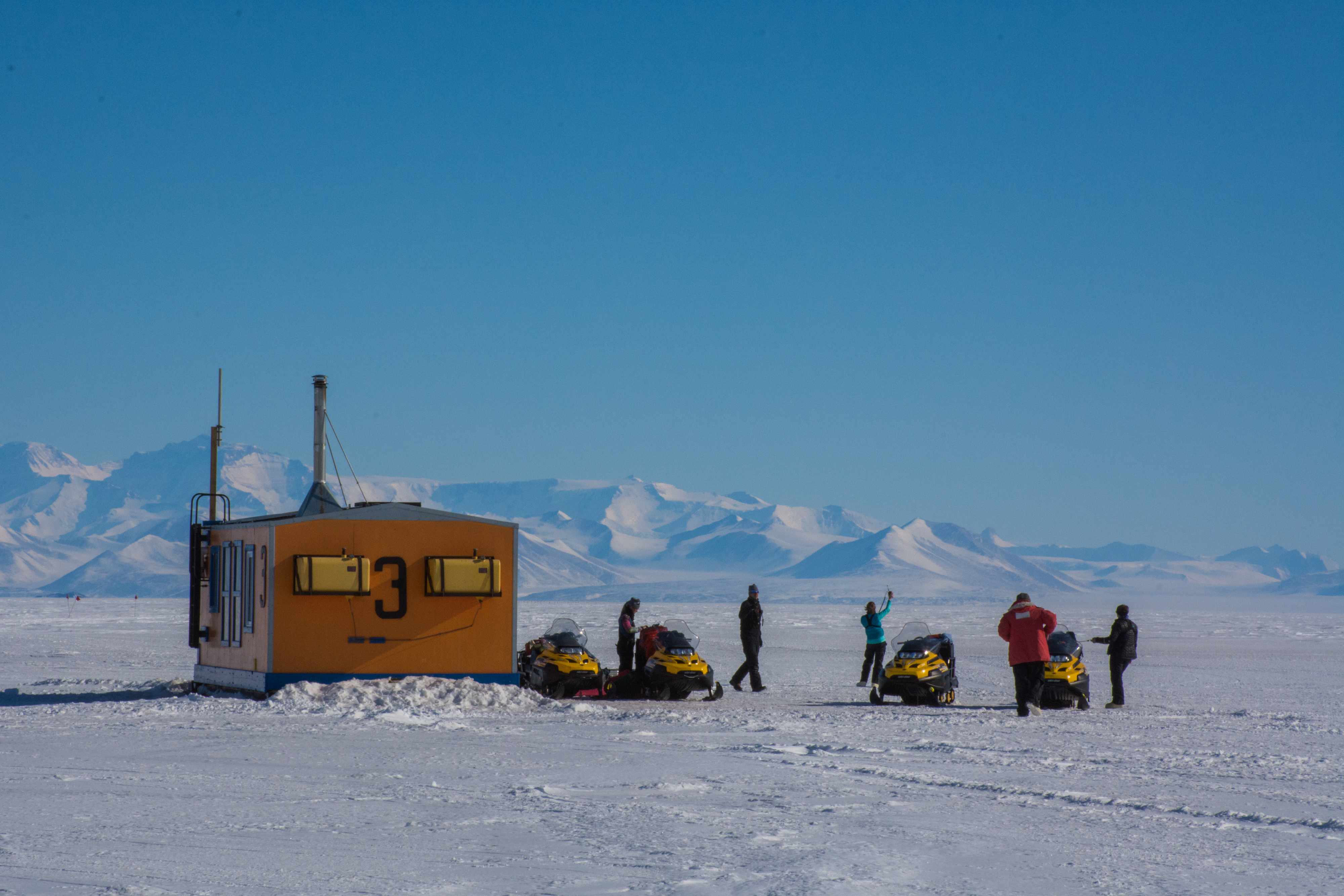 Four people next to four snowmobiles and a dive shack. 