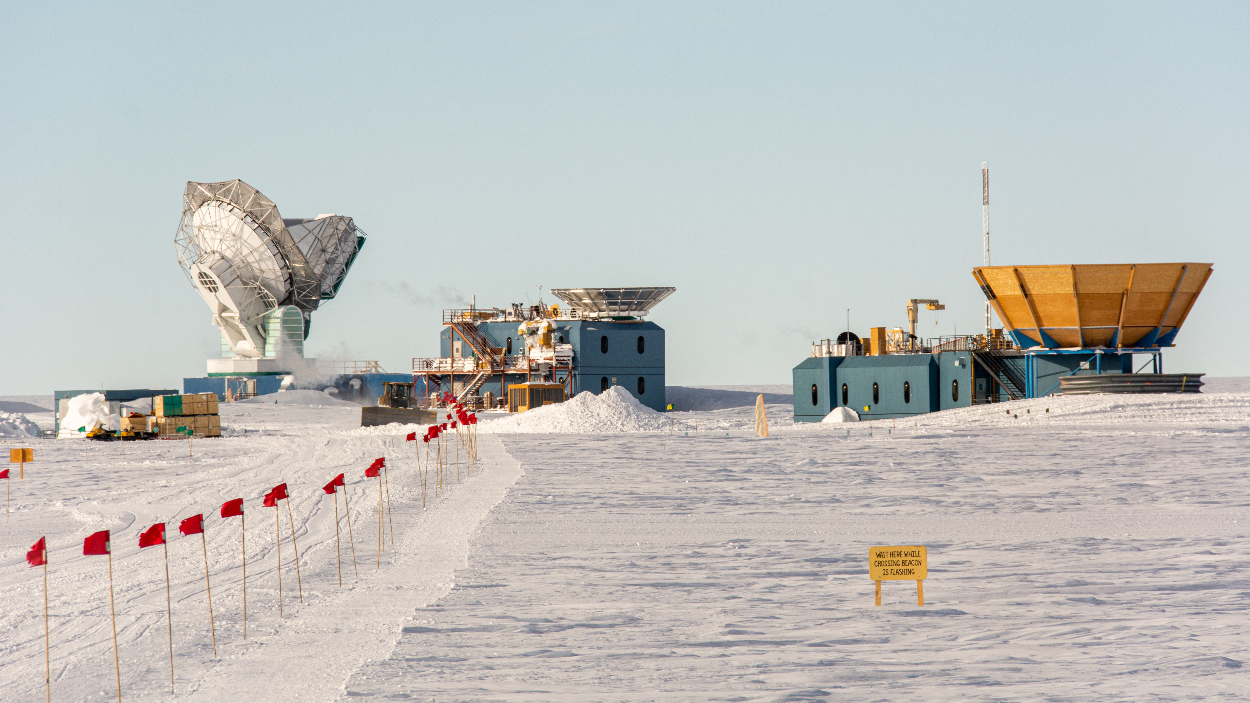 A line of red flags leads to far off buildings and a radio telescope amidst a snowy landscape. 