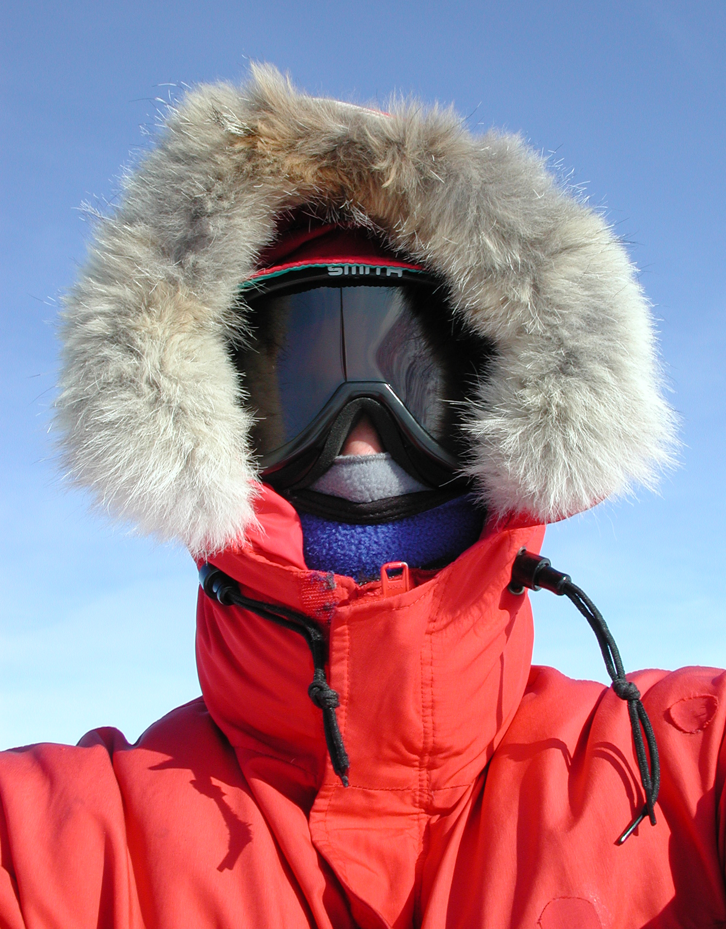 The head and shoulders of a person fully covered with goggles and red parka hood.