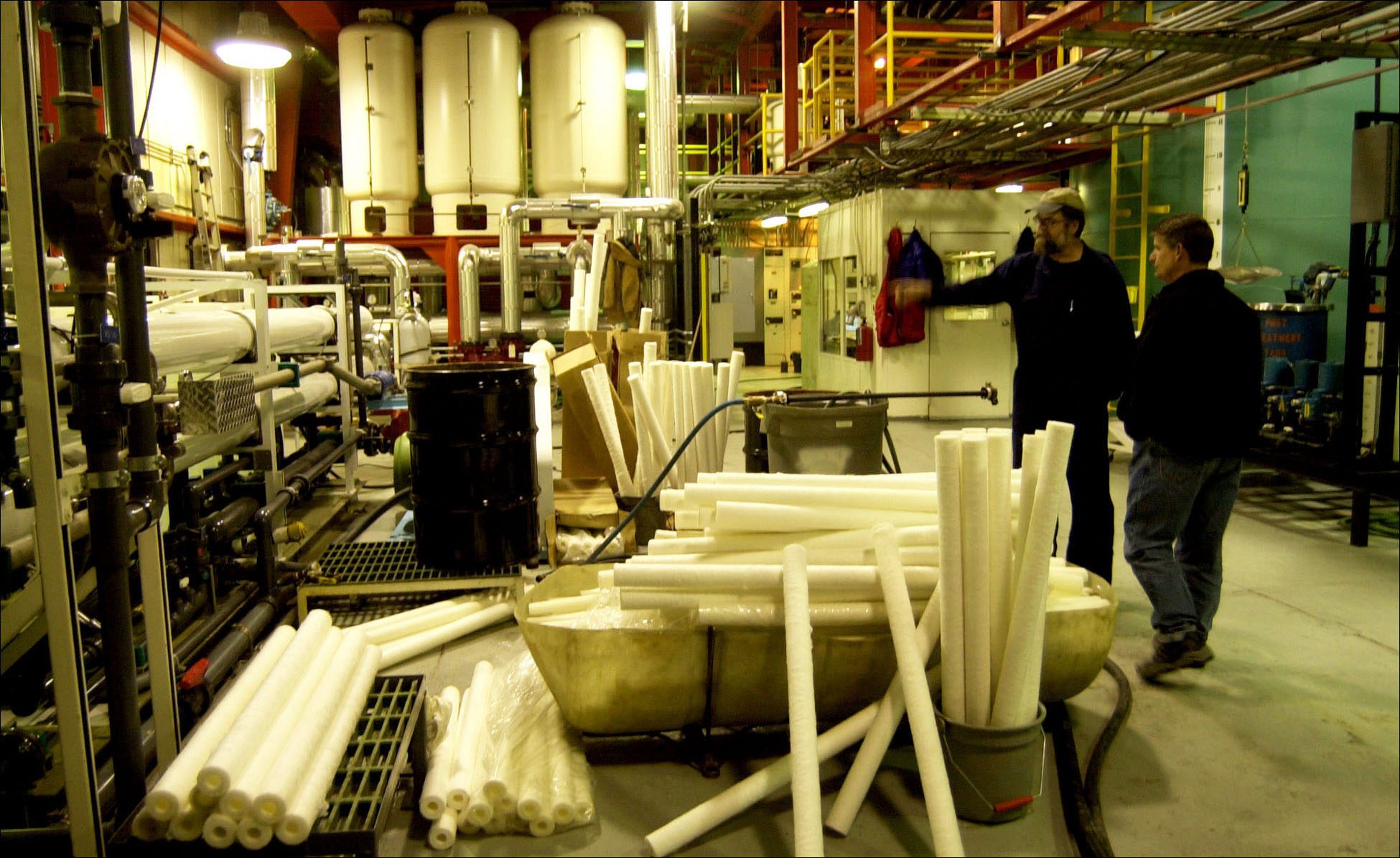 Two men look stand in a room with looking at dozens of long white tubes. 