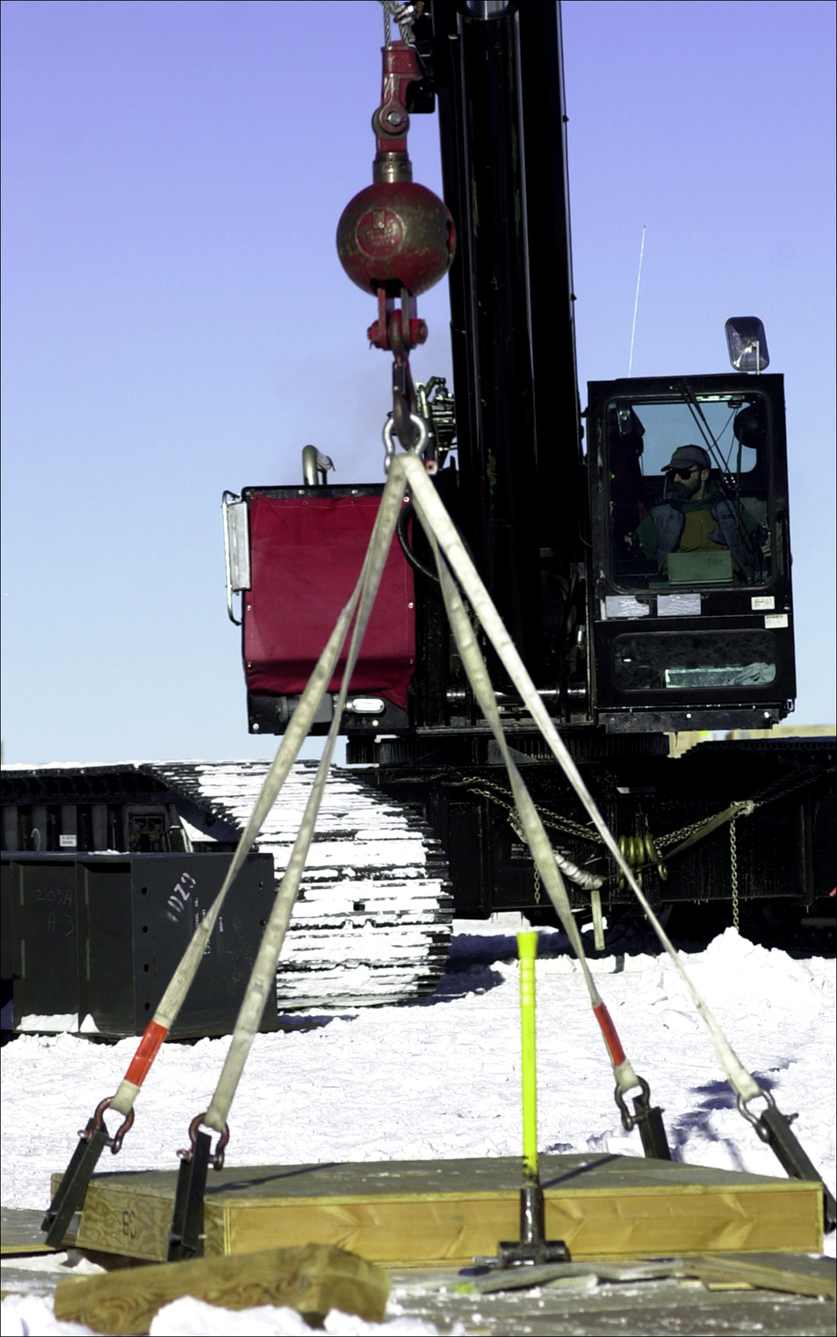 A black crane places a wood platform on the snow.