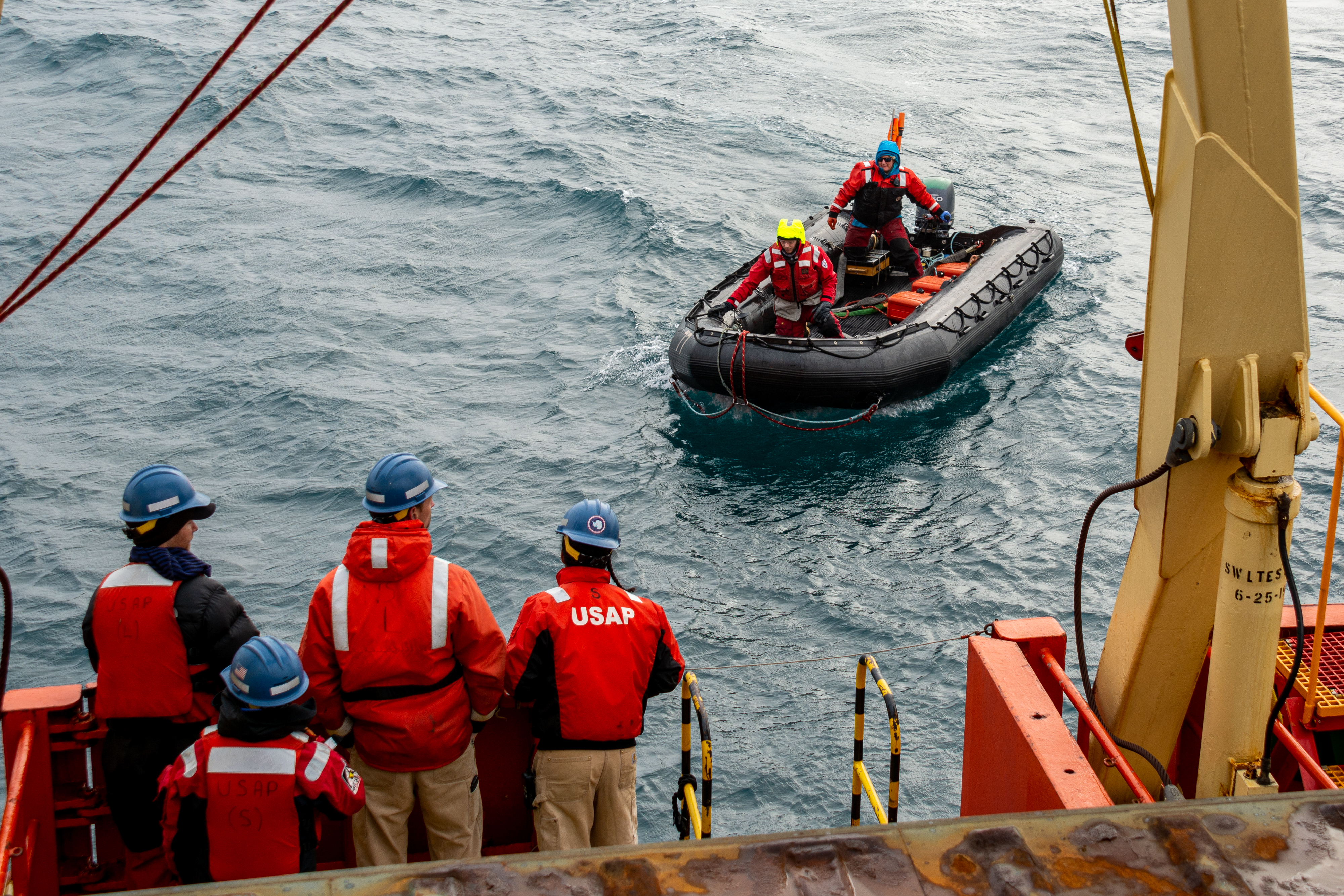 A small inflatable boat with two people navigates towards a ship with people on deck. 