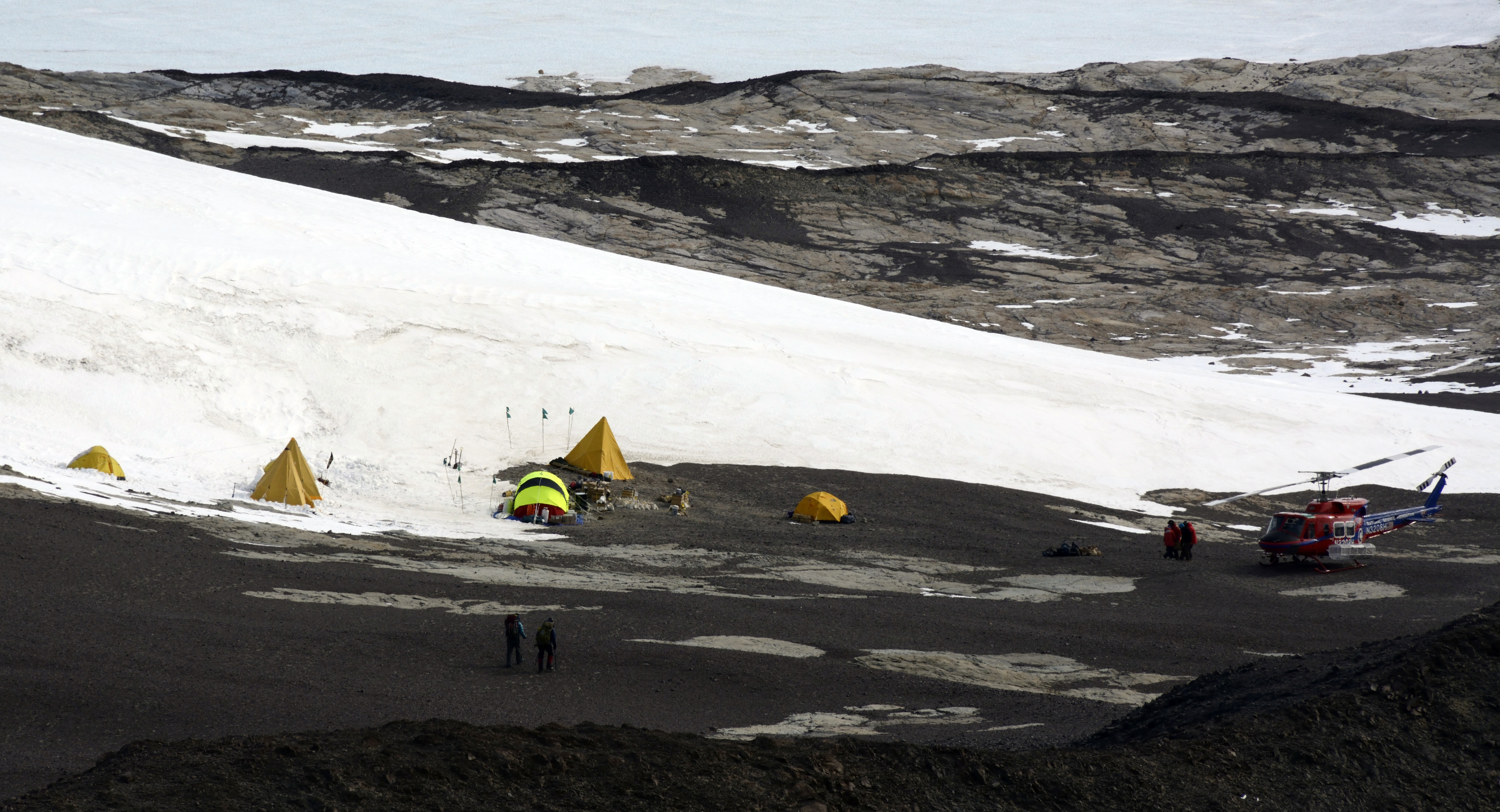 Helicopter sits on ground next to small camp and ice field.