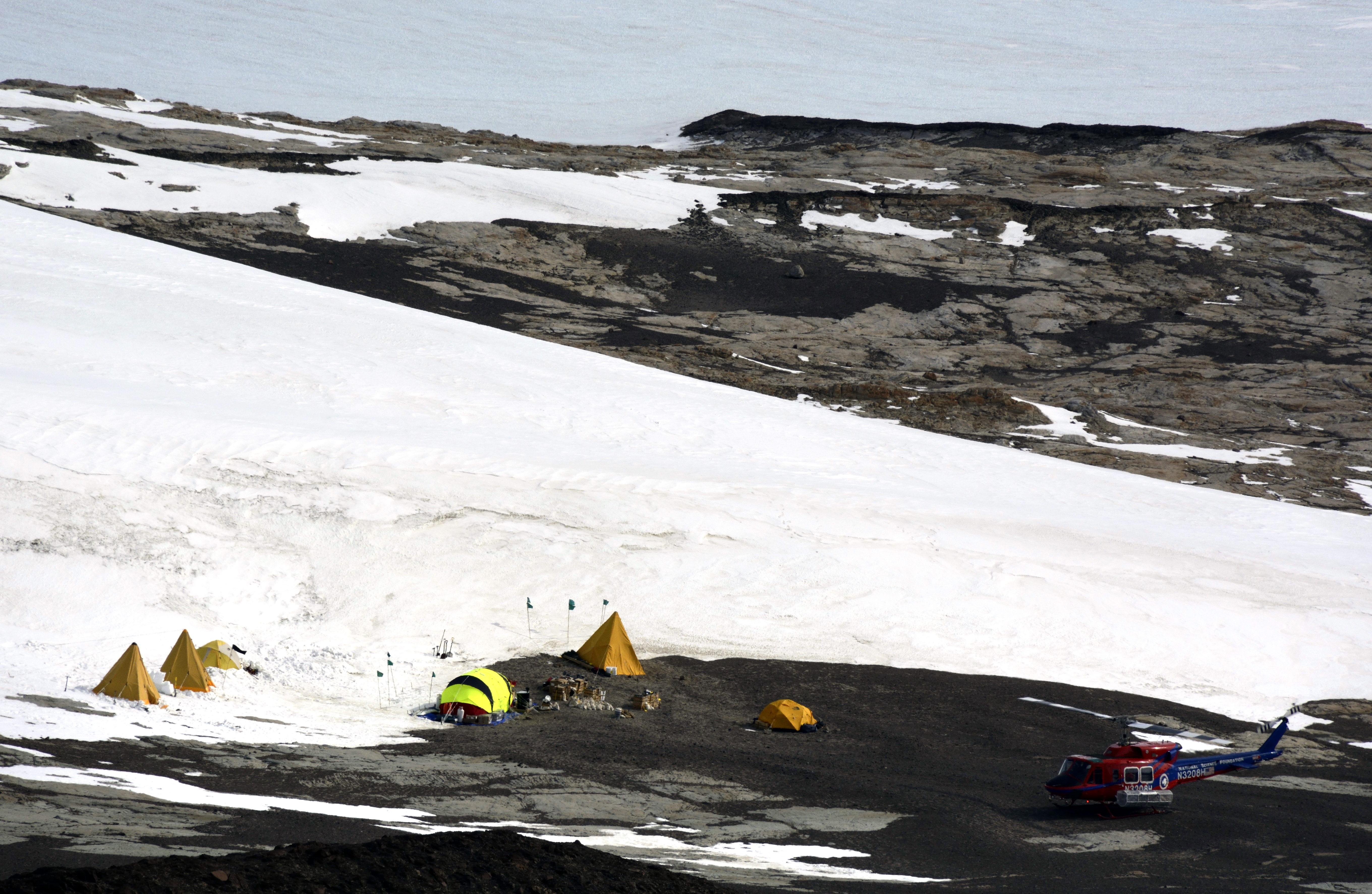 Helicopter sits on ground next to small camp and ice field.