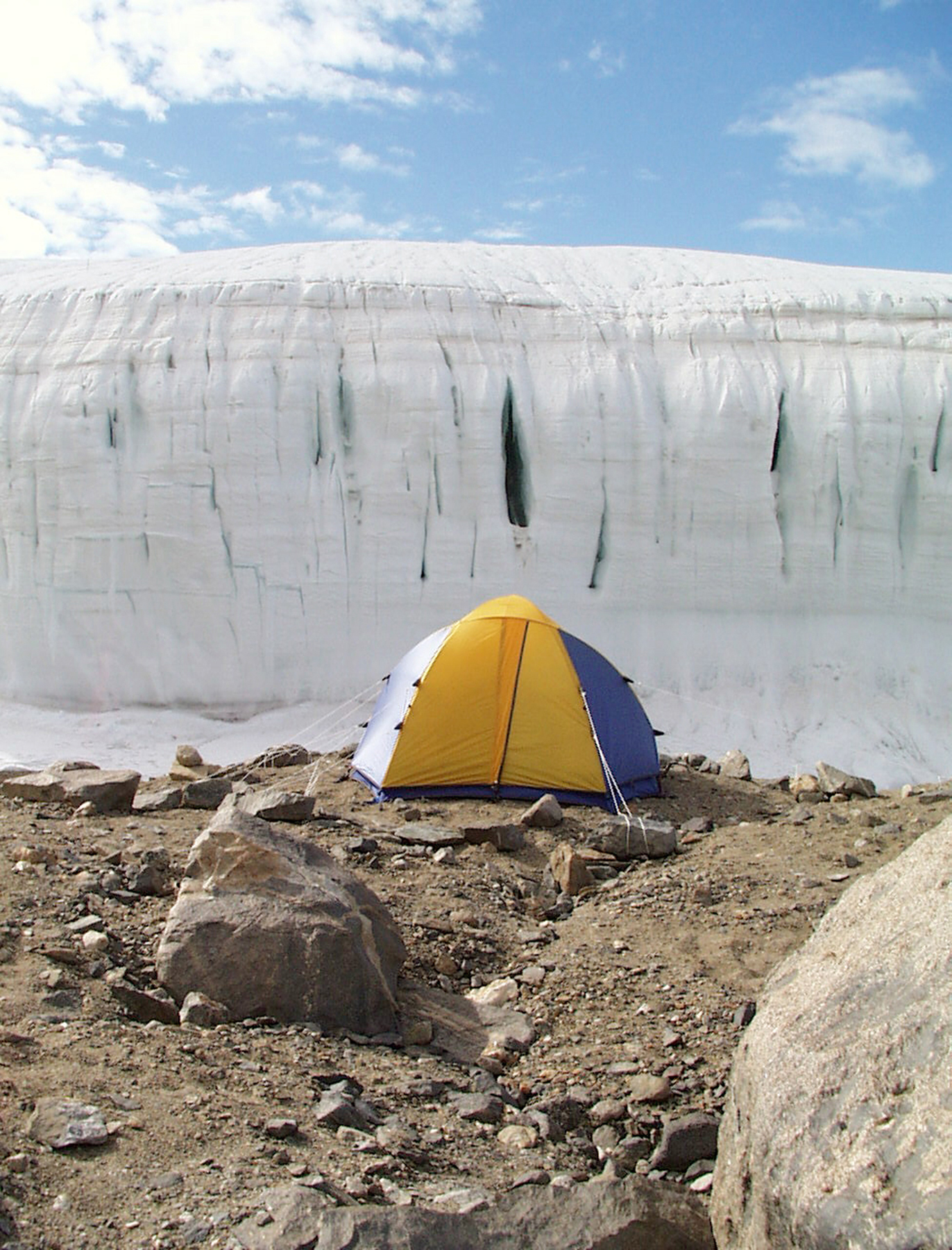 A yellow and blue dome tent sits in front of a glacier.