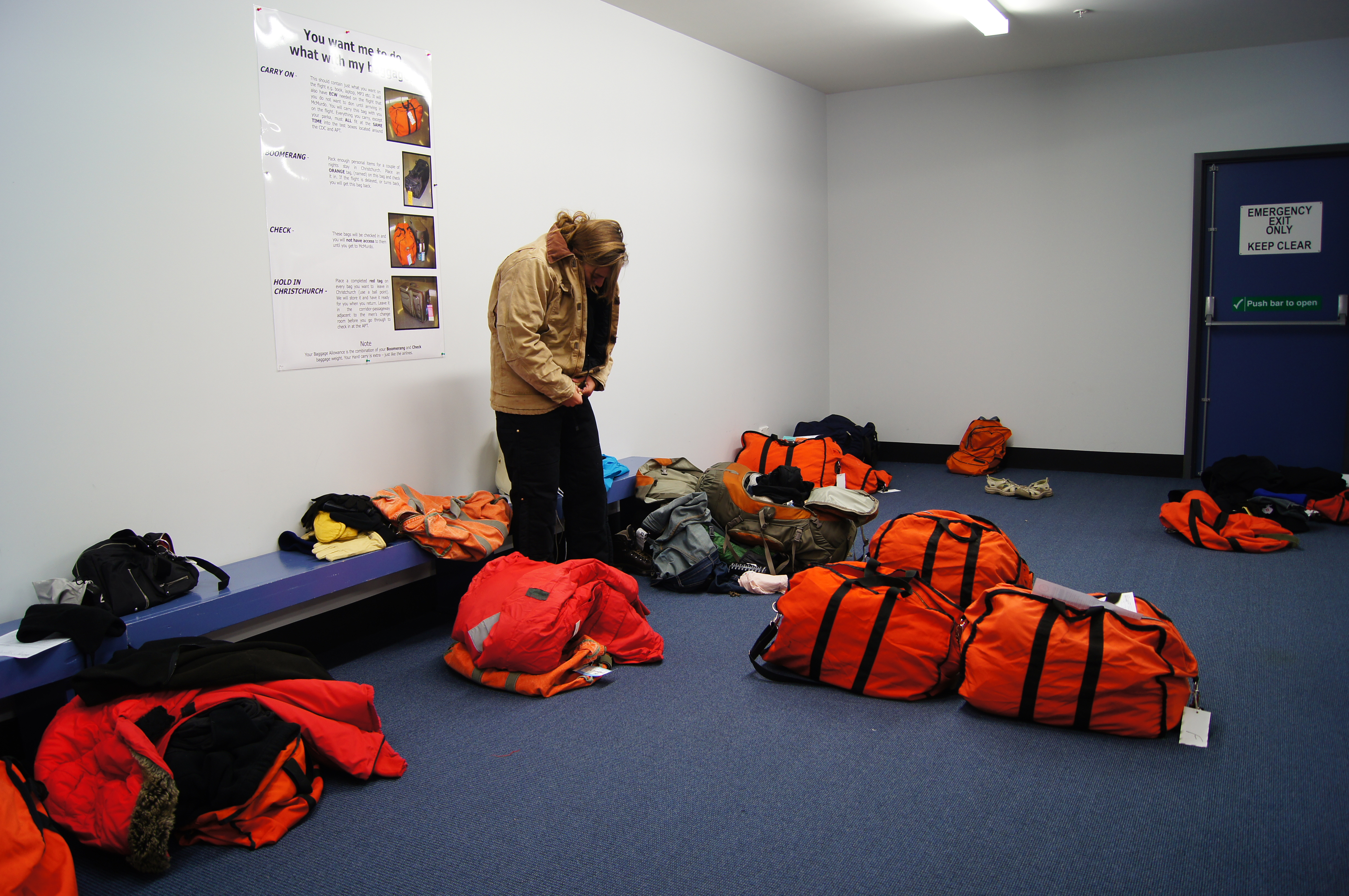 A woman tries on a coat in a dressing room littered with large orange duffle bags.