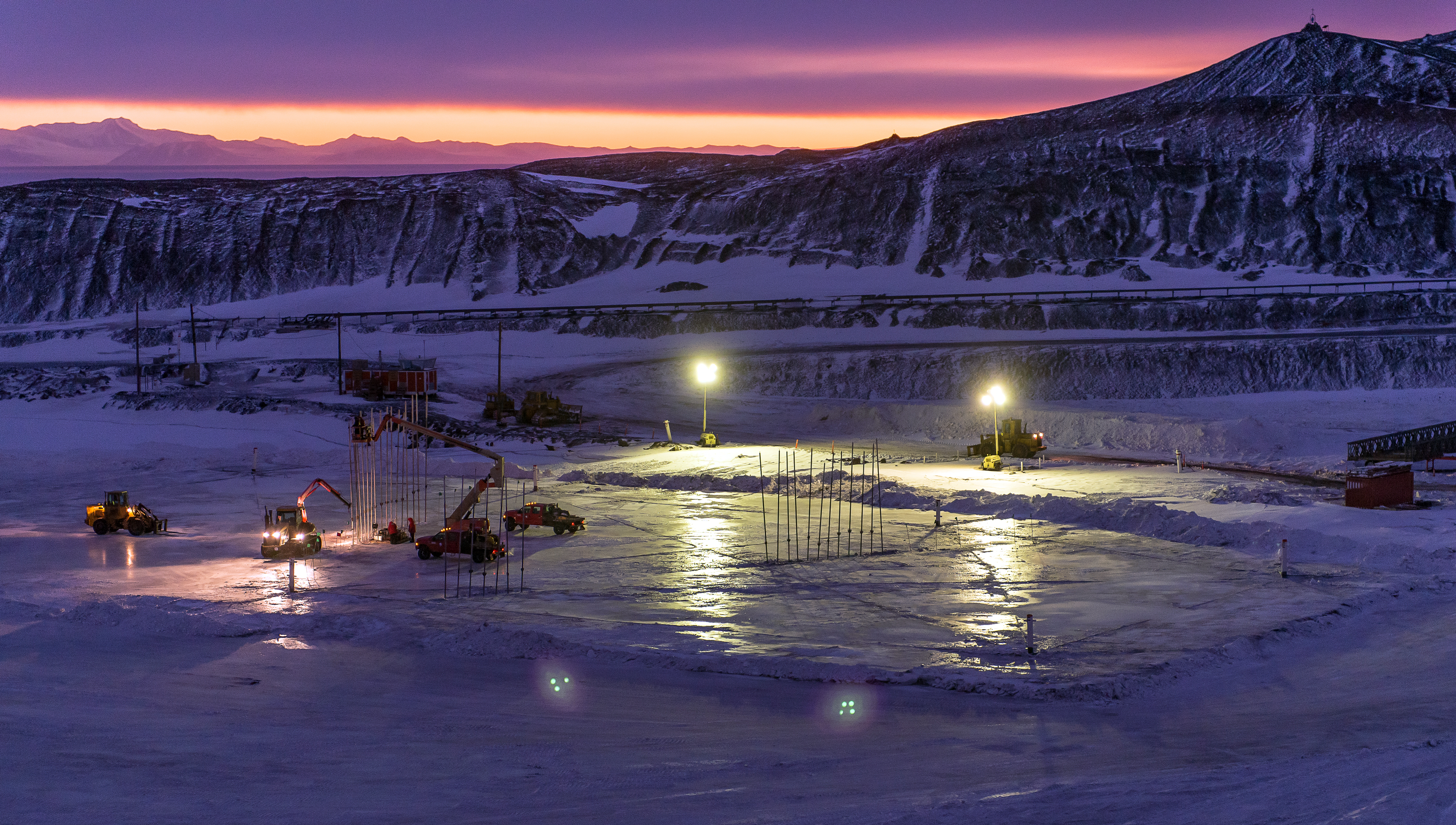 People and equipment work on flat, icy area at night.