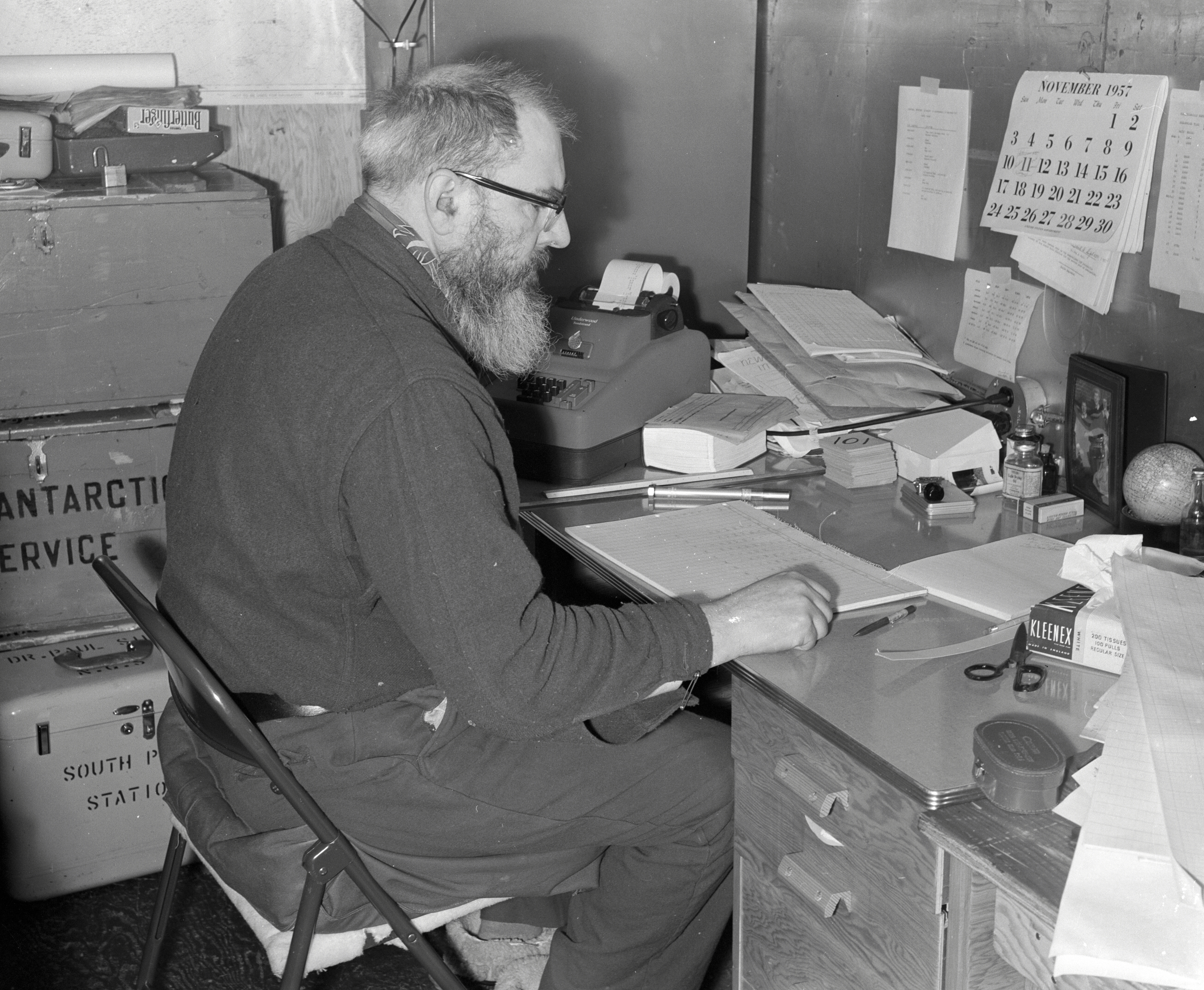 A bearded man works at a desk in November 1957.